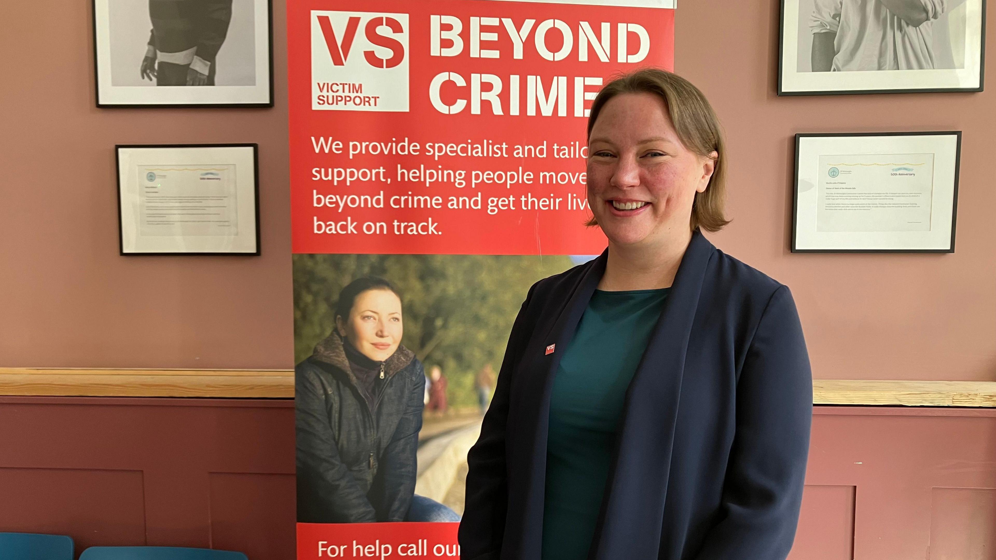 Katie Kempen wearing a green top and navy blazer standing in front of a red banner which says: "Victim Support, beyond crime". She is smiling at the camera and is wearing a victim support pin on her lapel. 