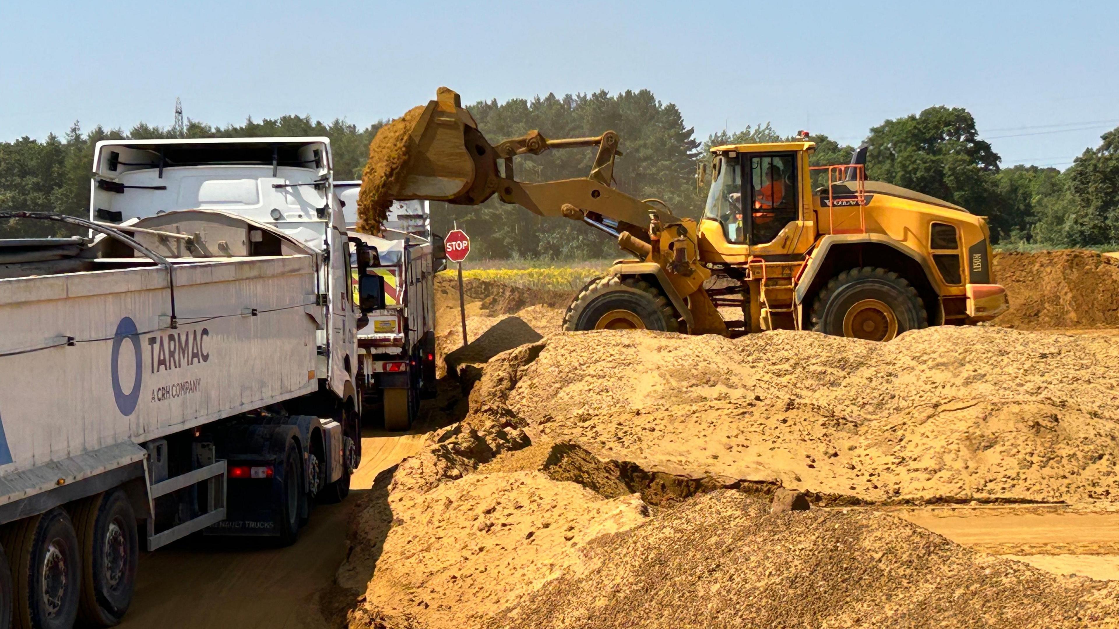 A yellow digger on the right is dropping sand into a large which truck to the left, surrounded by a sandy quarry.