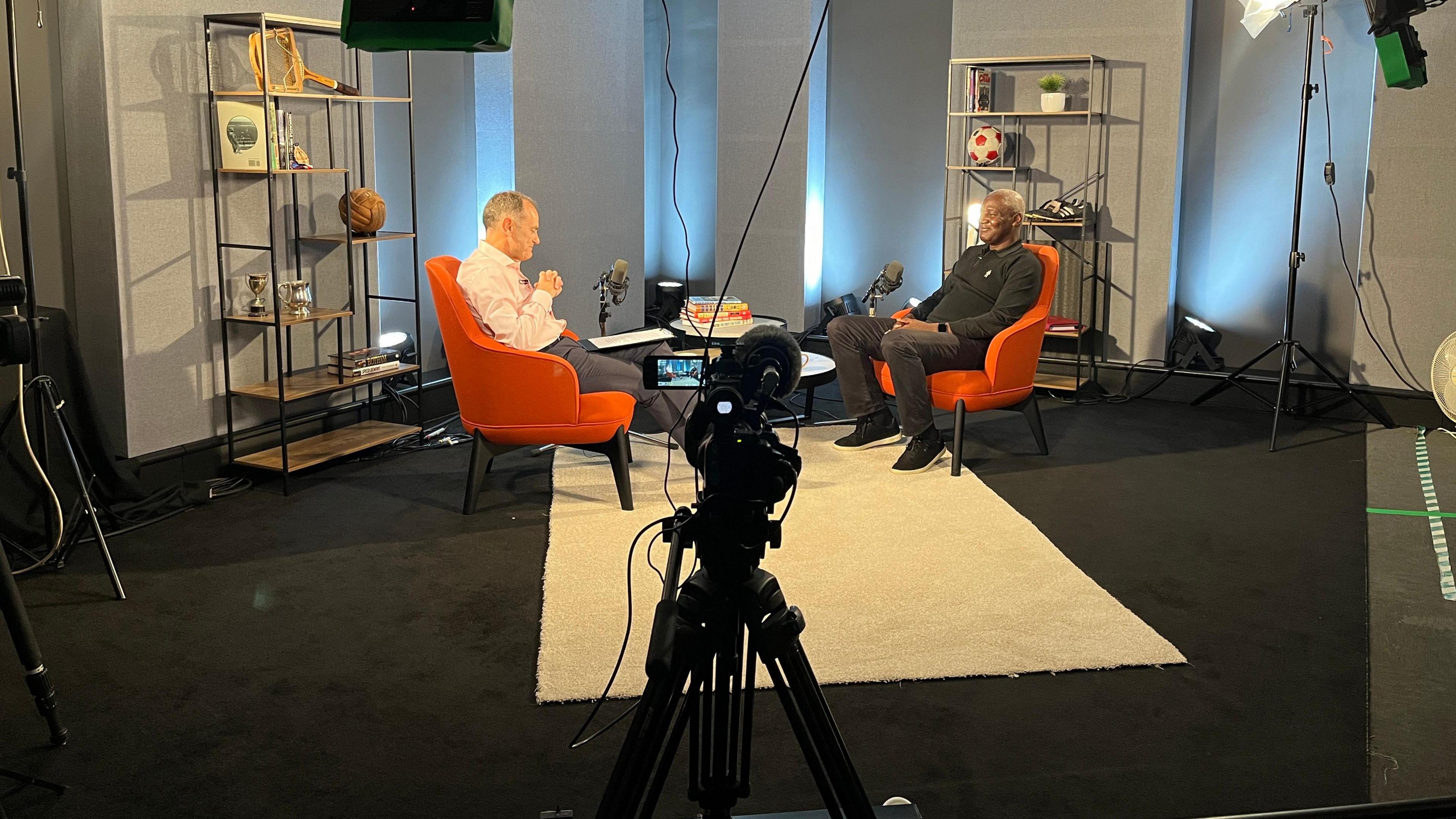 A behind the scenes shot of Jeff Brown, in pale pink shirt and grey trousers, talking to Gary Bennett, who is wearing a black jumper ad dark grey jeans. In the foreground, there is a microphone and a camera placed on a tripod. 