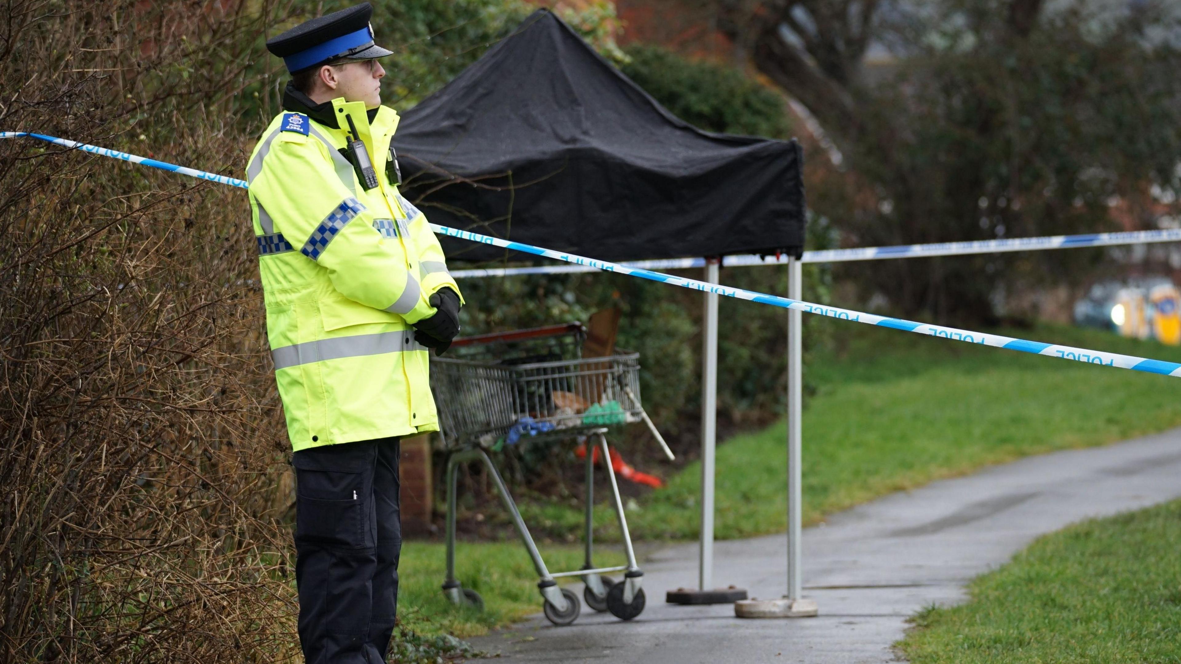 A police officer stood beside the cordoned scene of an attempted murder in Eastbourne, East Sussex. A trolley can be seen in the background under a black canvas. 