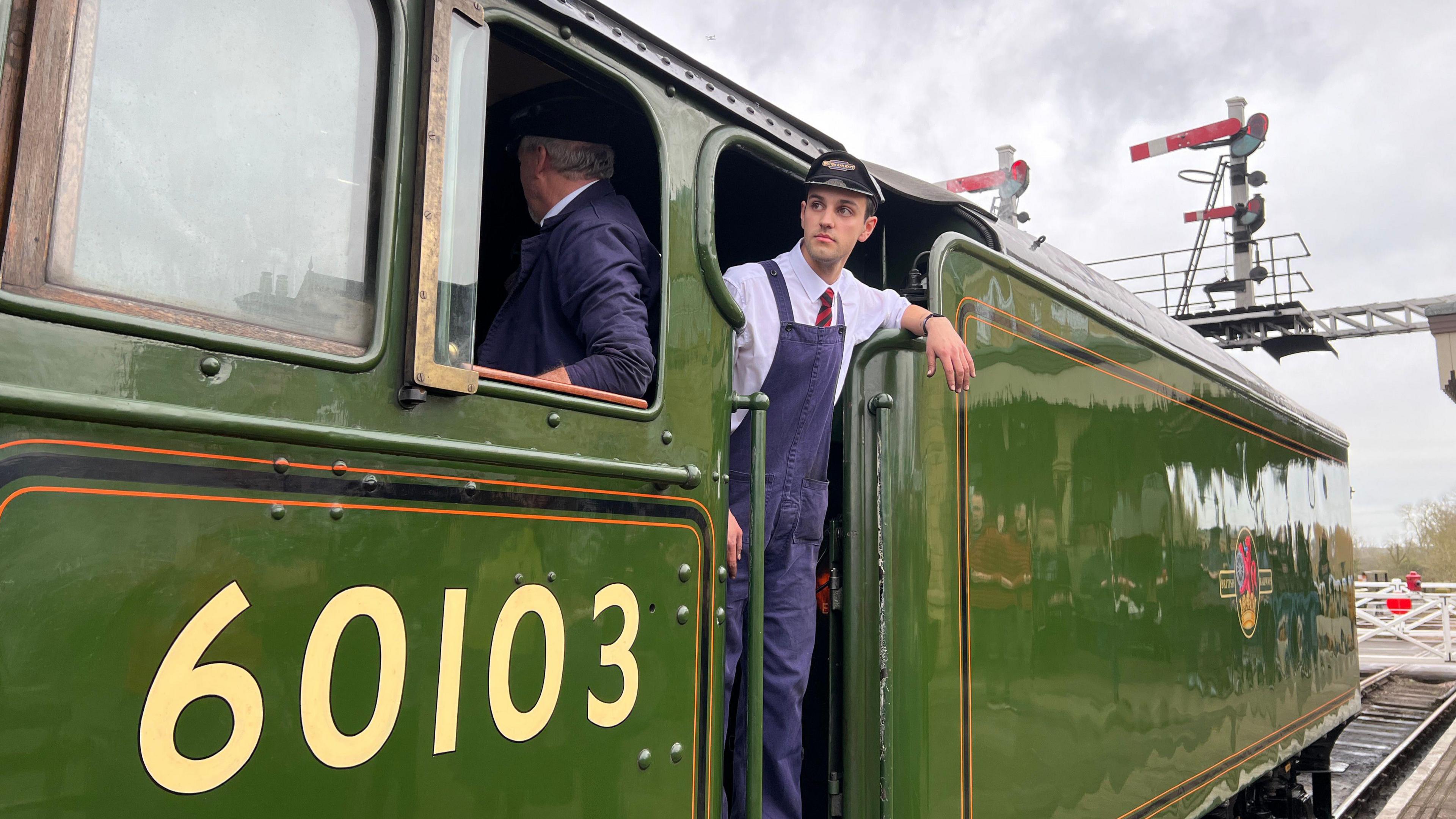 A rail engineer wearing blue dunagrees leans out of the Flying Scotsman and loks along the platform. The steam engine is painted dark green. It has the number 60103 on its side.