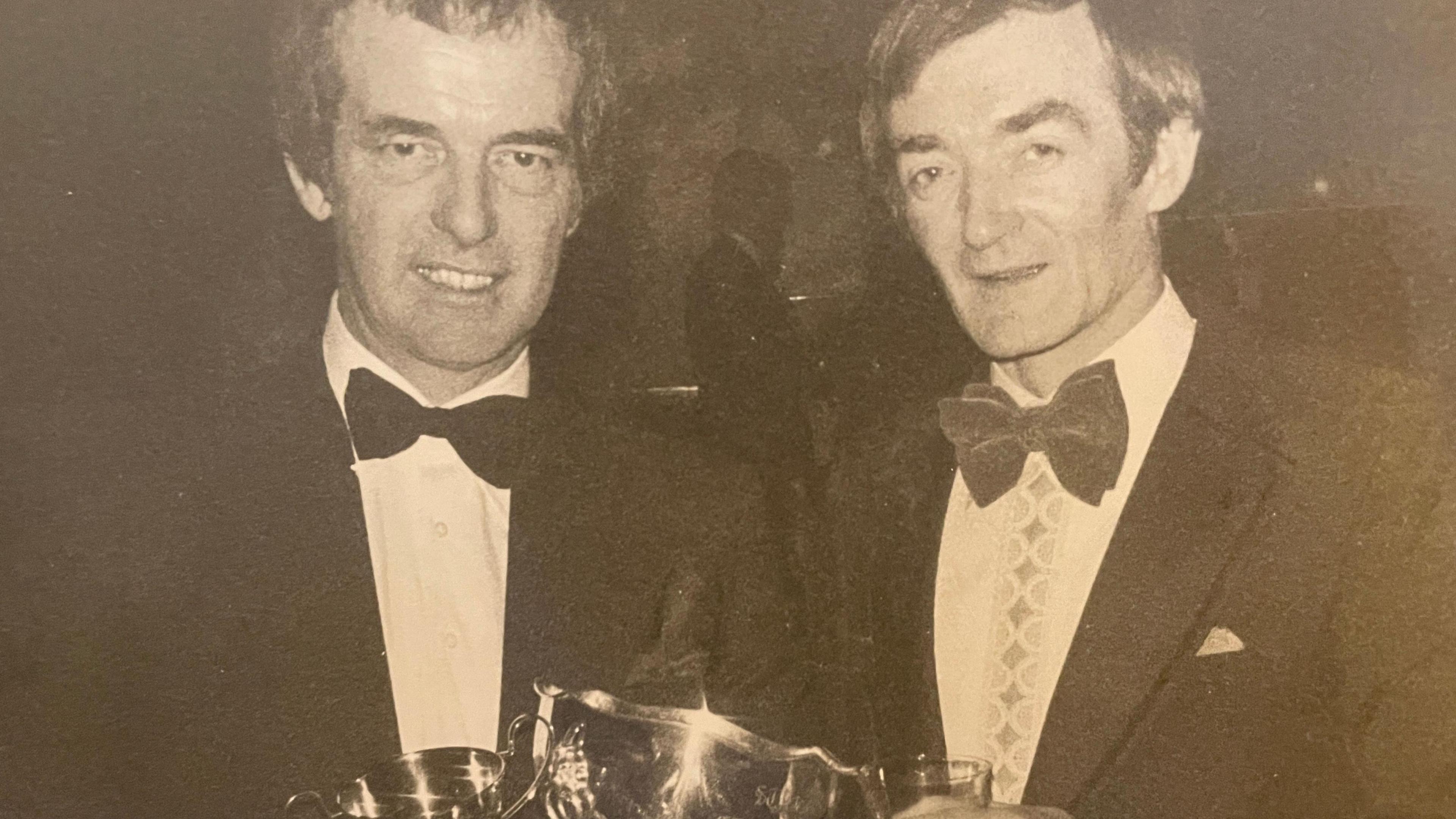 A black and white photograph of Roz's father Ron, he is on the left hand side of the frame. Ron is holding a silver award cup which glistens from the light of the camera's flash.