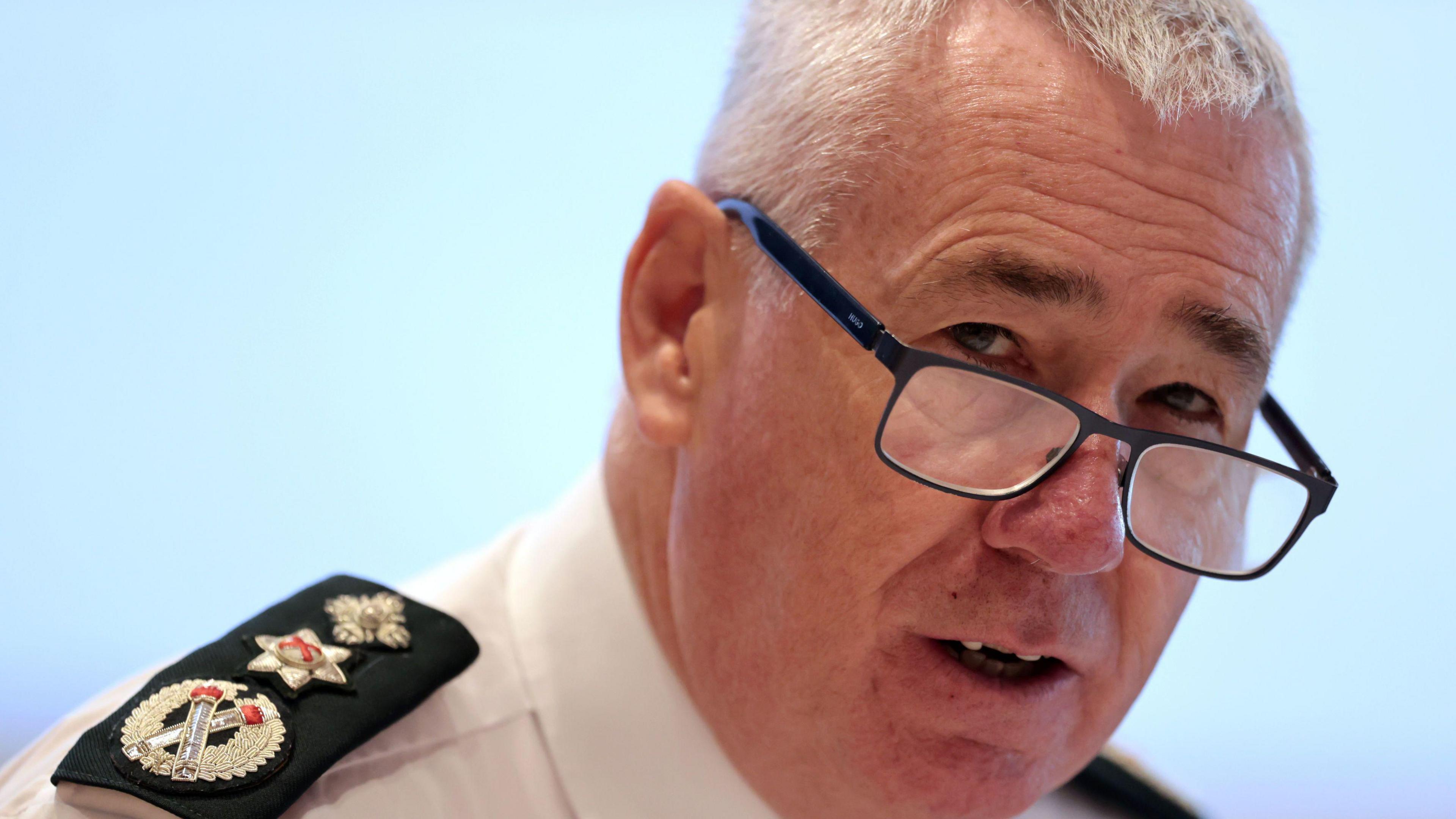 Jon Boutcher has short grey hair and is wearing rectangular black glasses, as well as a shirt with black badges on his shoulders.