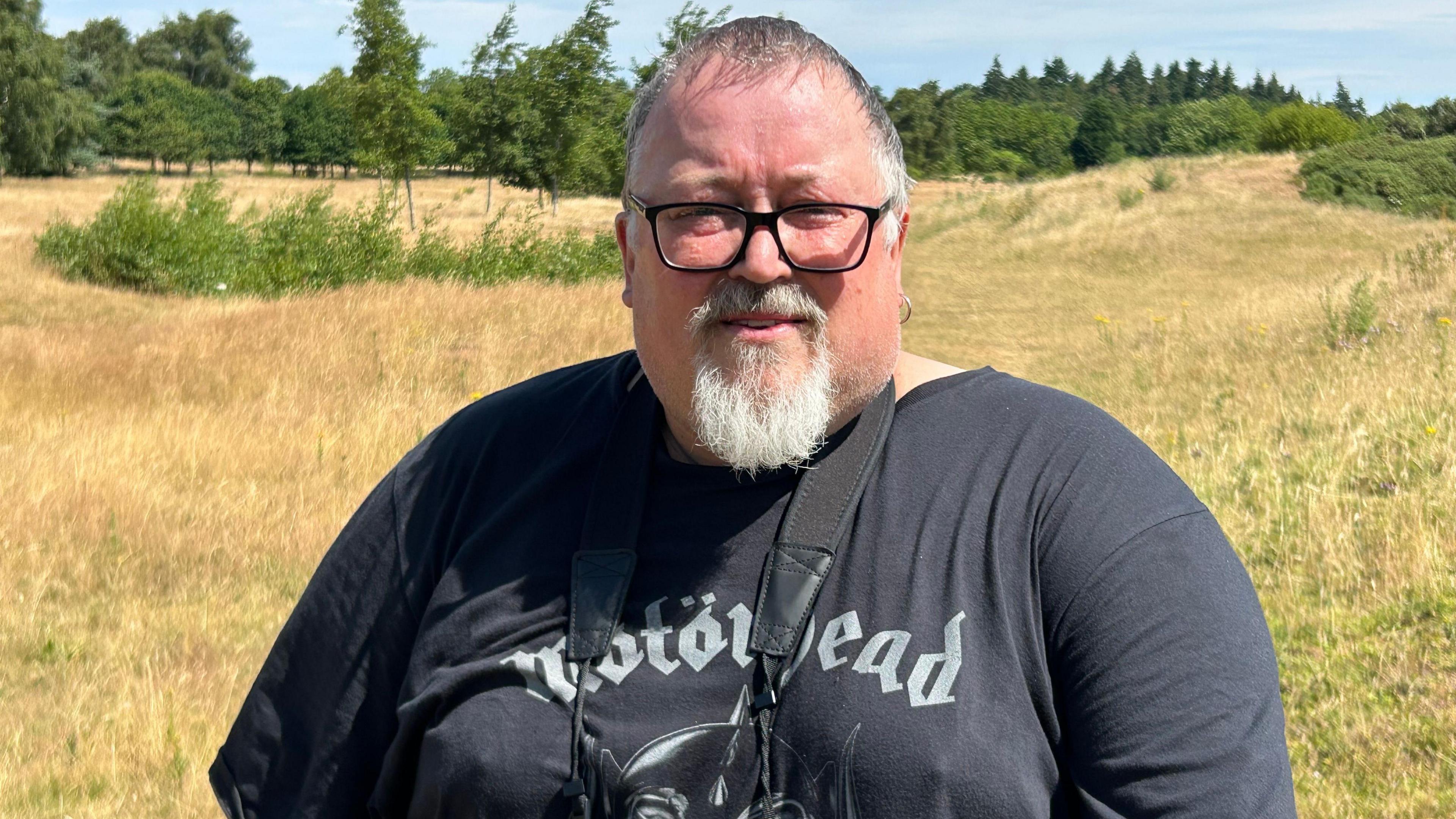 A man with short grey hair, a white goatee and black-rimmed glasses standing on a patch of grassland on a sunny day. Many trees are in the distance. He is wearing a black Motorhead t-shirt and has a pair of binoculars hanging around his neck.