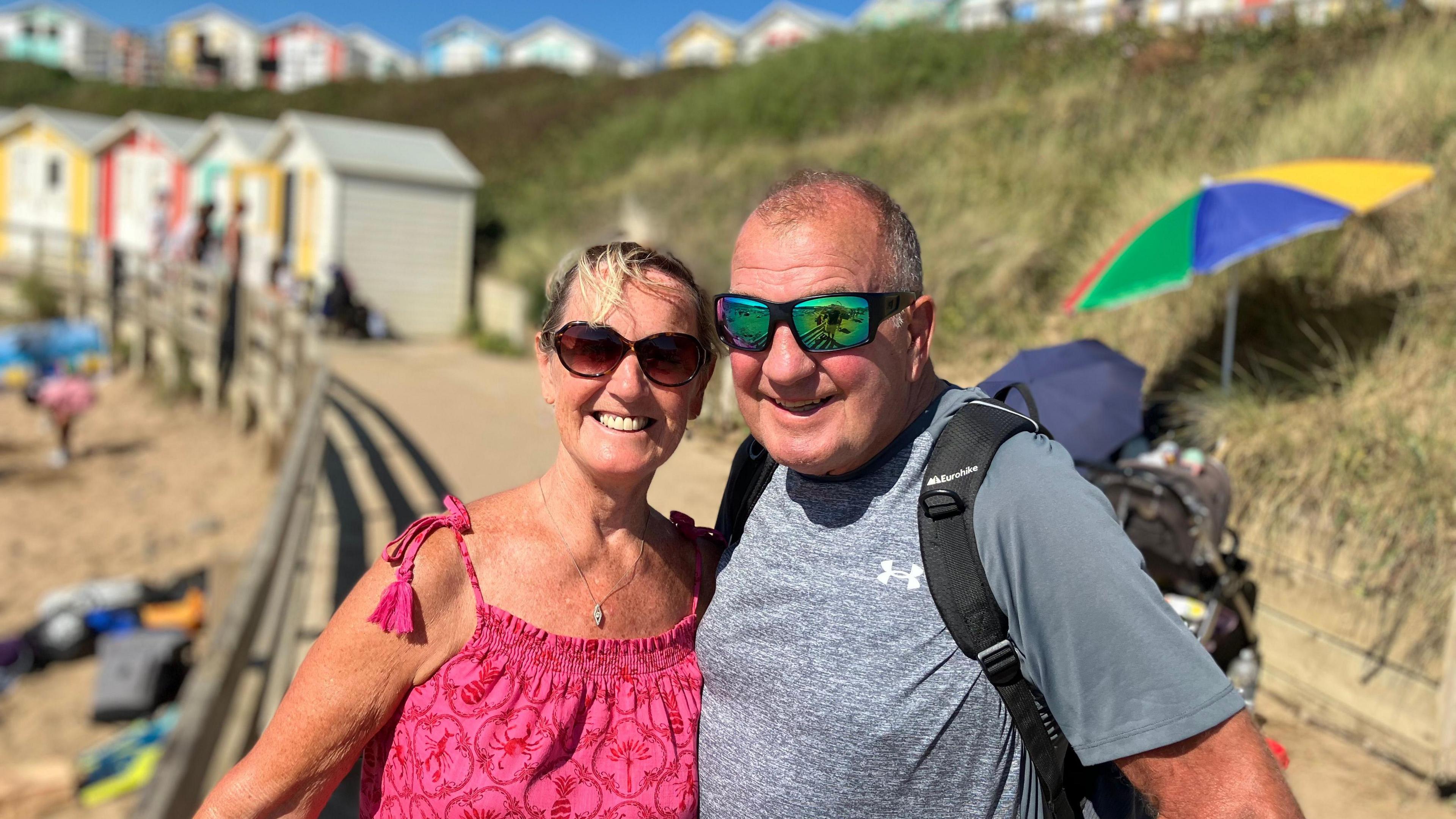A couple stand on a raised walkway, next to a beach. The sun is shining and they are standing in front of colourful beach huts 