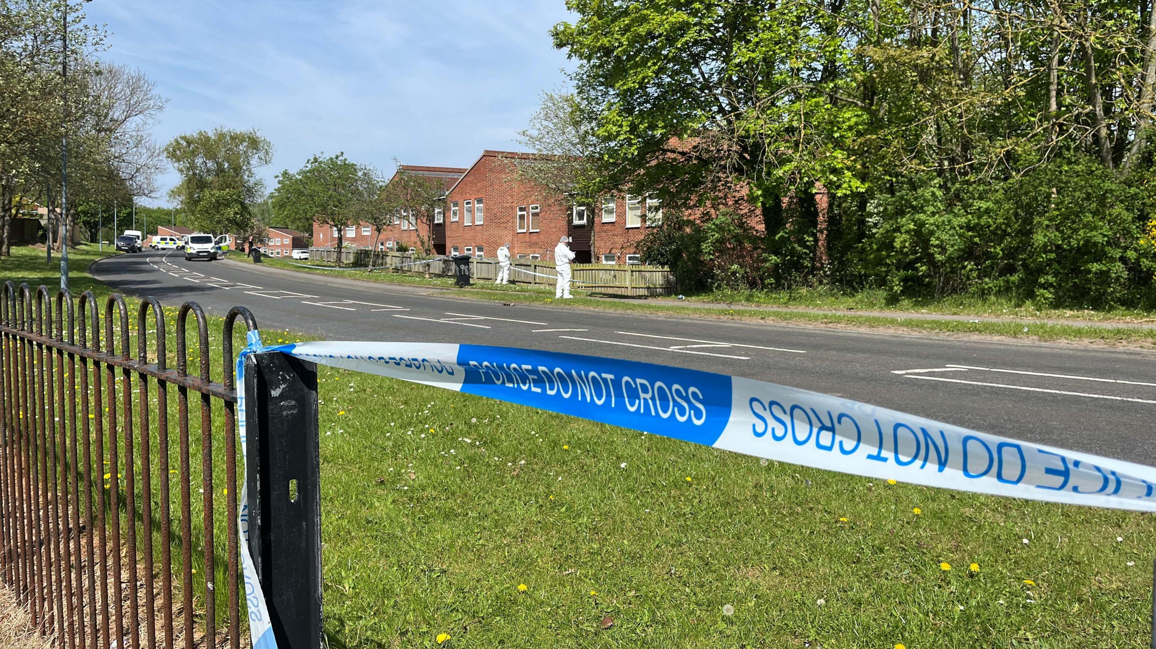 In the foreground, blue and white police tape has been attached to a black fence. In the background, forensics officers in white suits are inspecting ground near a fence with a row of houses behind.