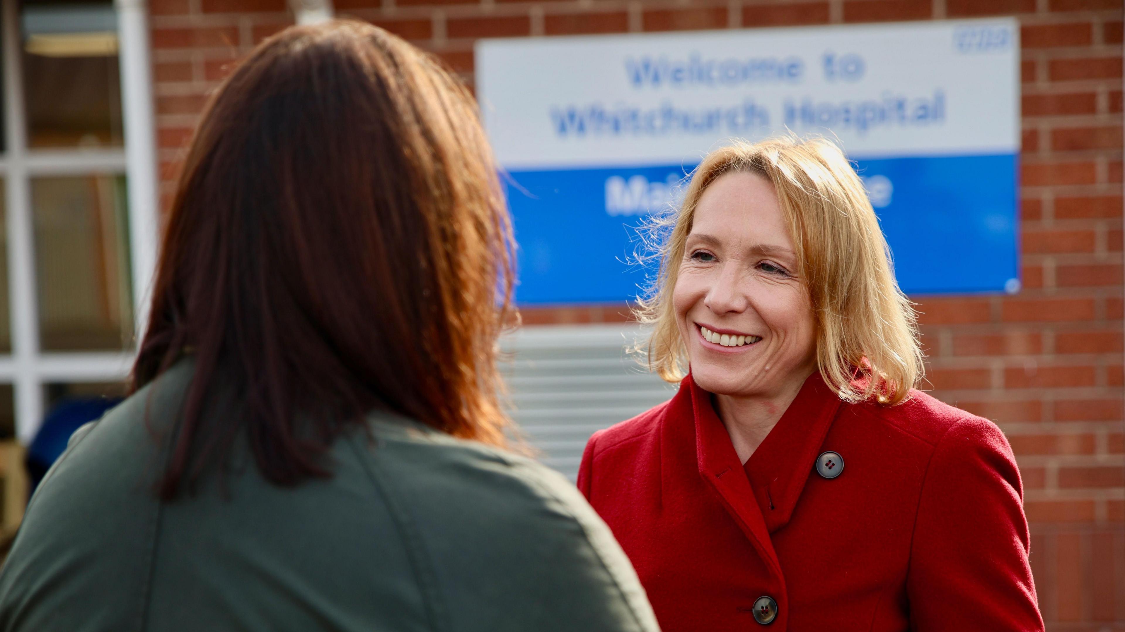 Helen Morgan MP is talking to a person who has their back to the camera. Ms Morgan is standing in front of a sign that says "Welcome to Whitchurch Hospital". She is wearing a red woollen coat and is smiling.  