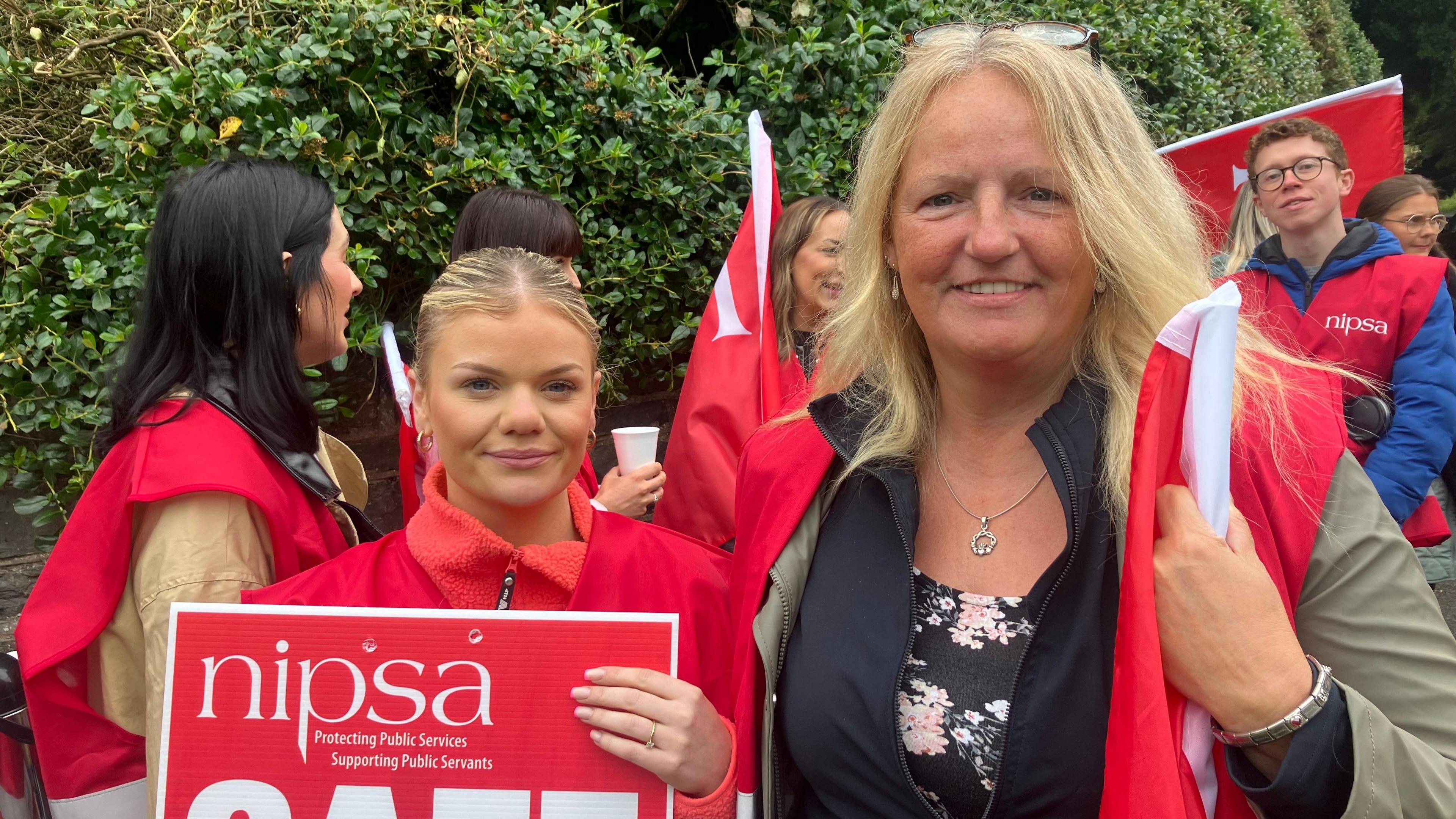 Two blonde women at a picket line with red flags and signs