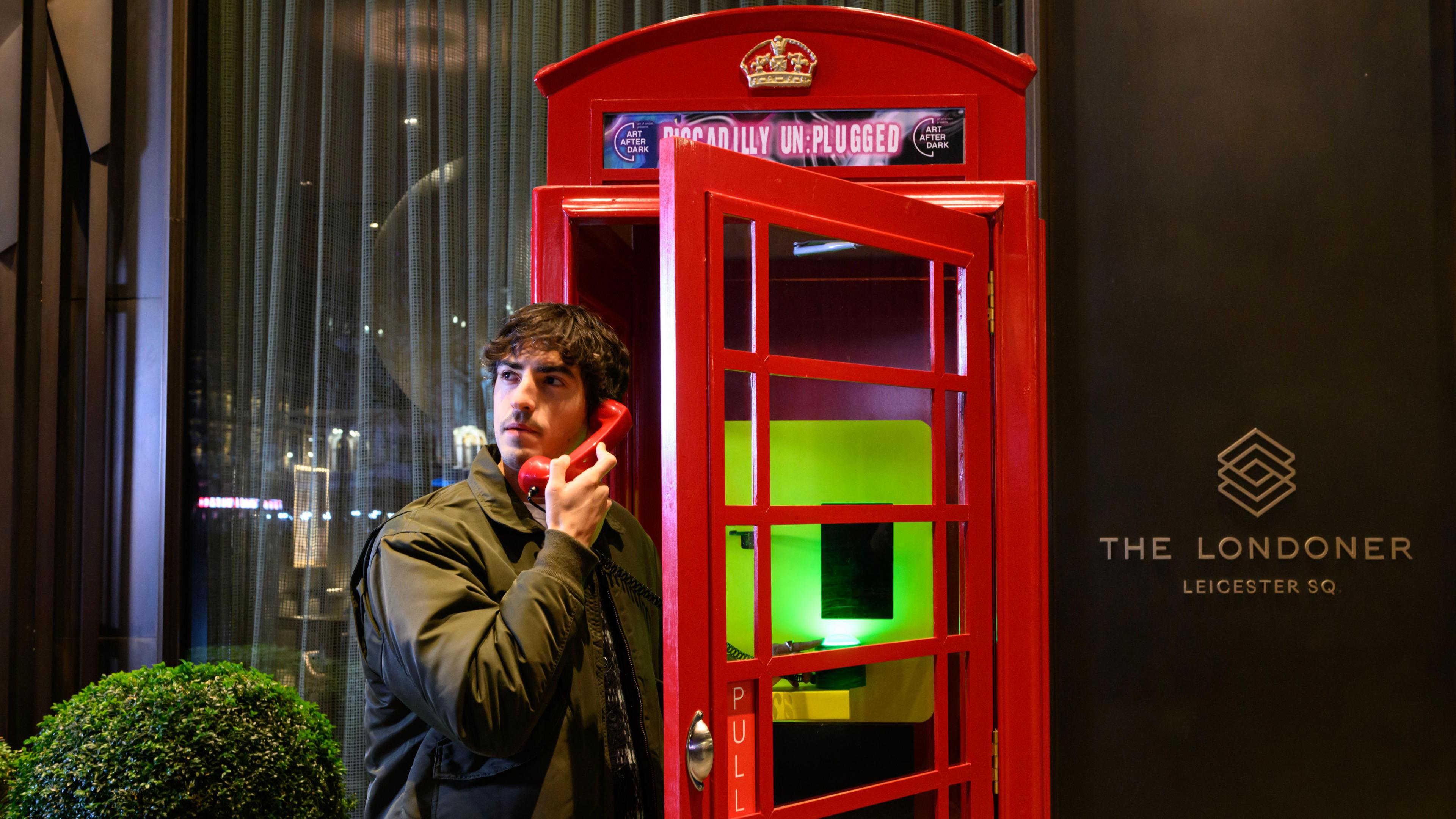 A man stands in the doorway of a red phone box. He is holding a phone and looking up to the right. Behind is a glass-fronted wall while to the front is a black wall with The Londoner brand written on.