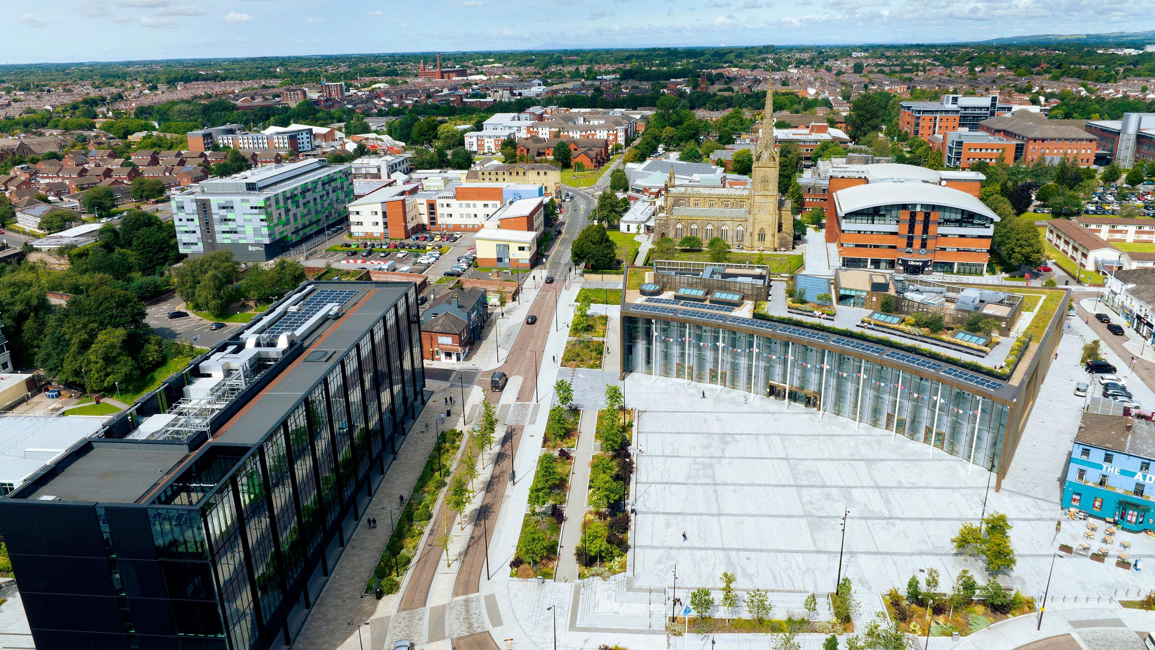 drone view of a modern multi-storey black building at the left of a pedestrian plaza with rows of shrubs and other multi-storey buildings and an old church in the background