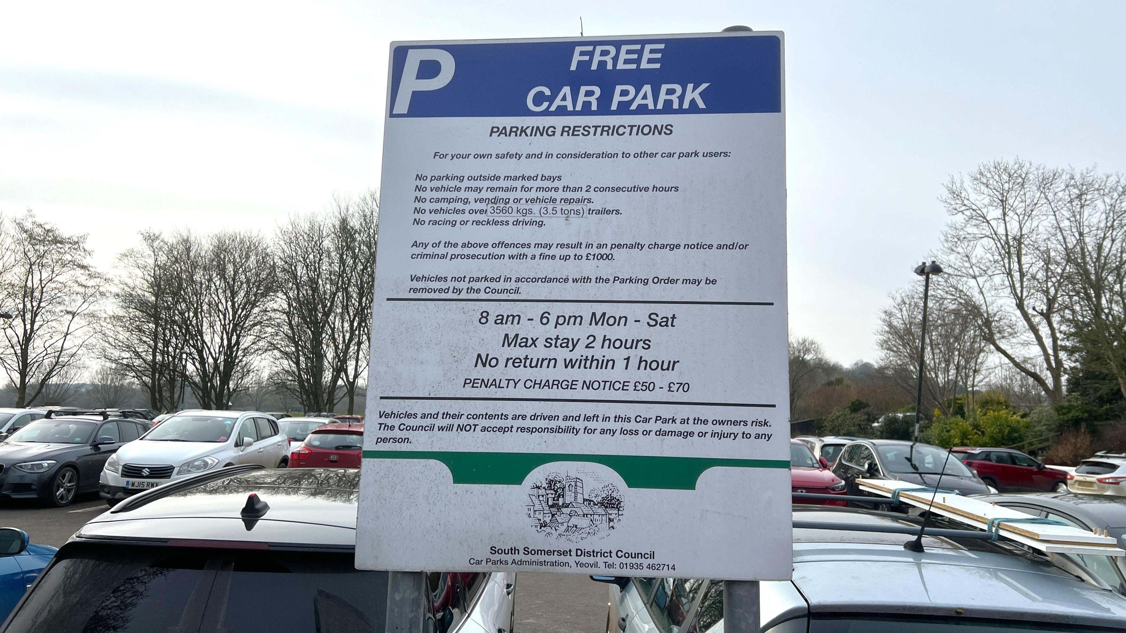 A blue and white car parking sign which says Free Car Park at the top. People can stay for two hours maximum. In the background the car park is completely full of cars and surrounded by trees.