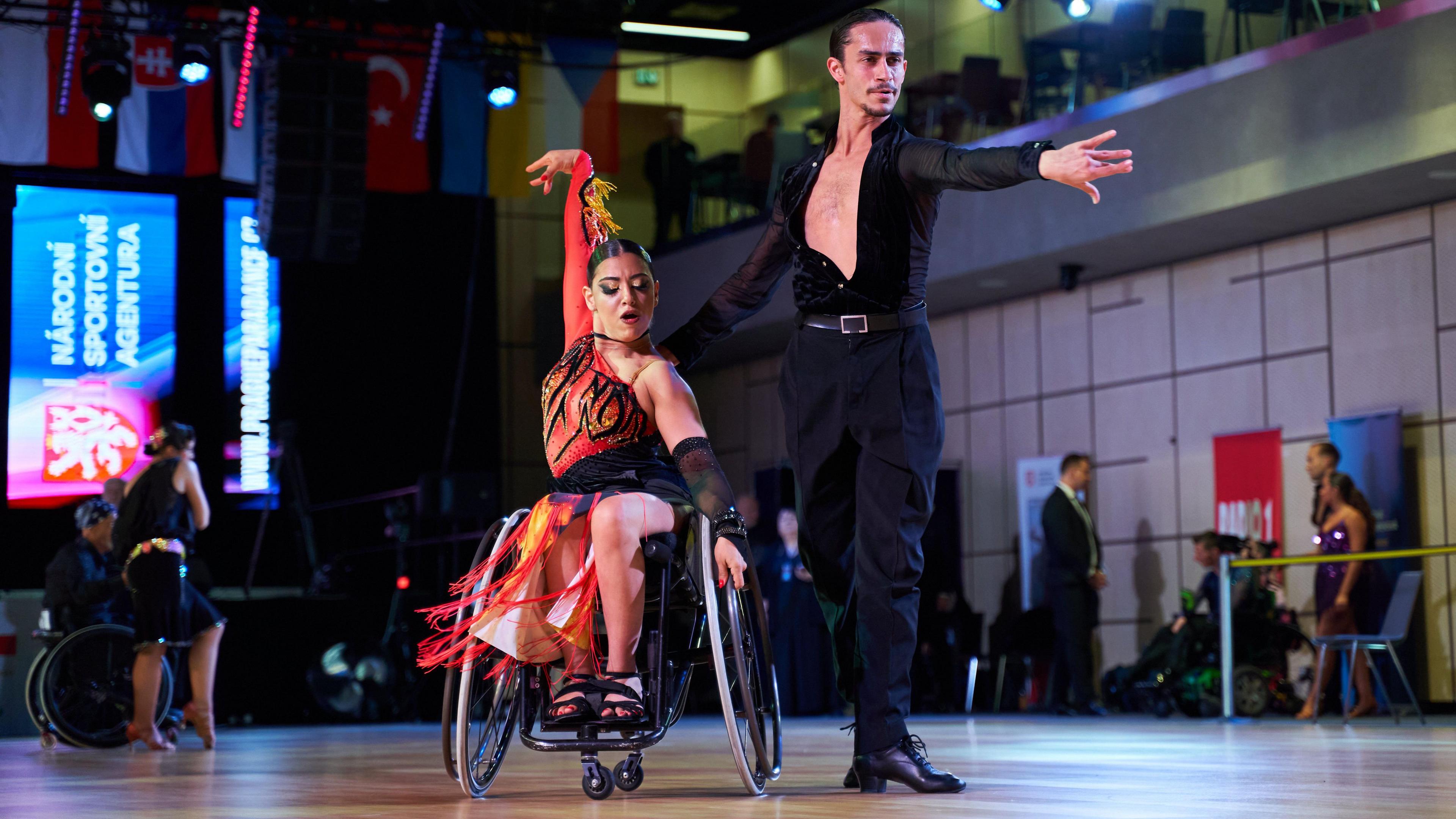 A woman in a wheelchair wearing a red fire constume dances with her arms in the air and her hair slicked back in a tight bun. Her dance partner is stood with his arms outstretched on the dance floor, he is wearing all black and looks focused.