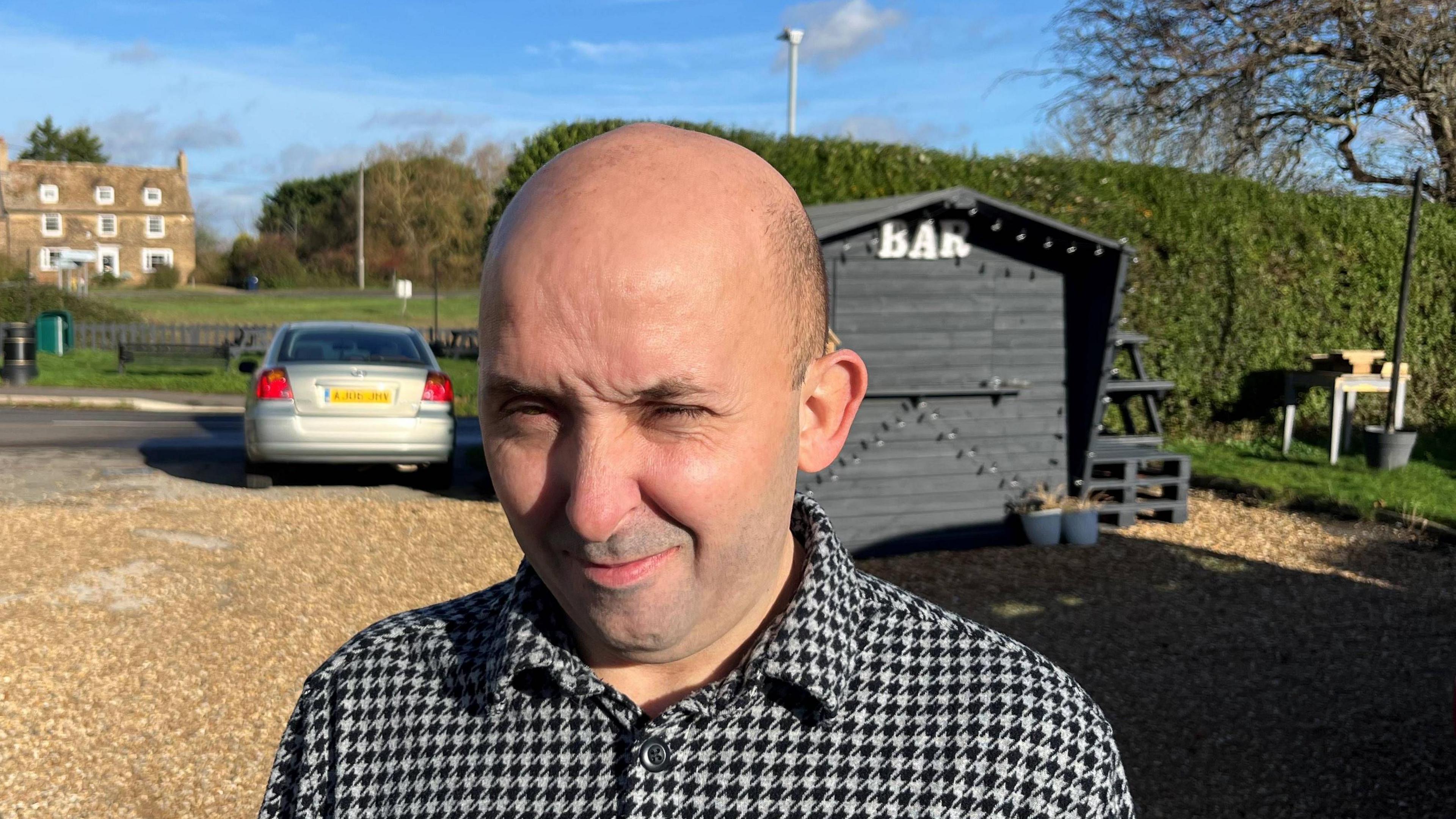 Brian Wadey, 40, has a bald head with some fine black hair on the sides. He is squinting a bit due to bright sunshine and is wearing a black and white patterned shirt. Behind him is a gravel driveway to his pub and a wooden shed with fairy lights strung up on it, and the word 'bar' on top. A tall hedge and a car can be seen too, with a large house visible in the background.