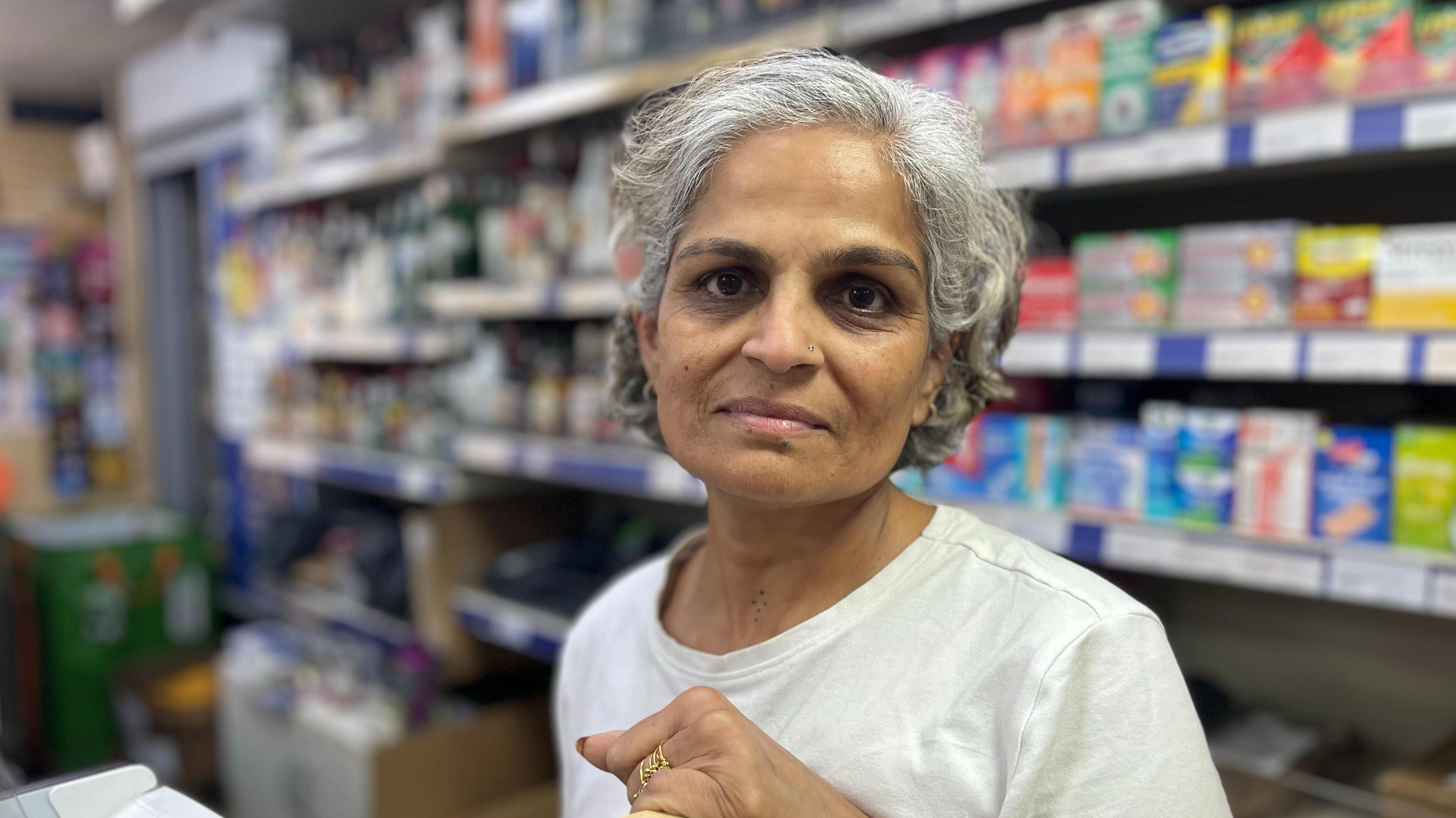 Neha Patel stands inside the Beach Shop near Shoreham seafront. She is wearing a white top and there are shelves filled with drinks, confectionery and everyday goods behind her.