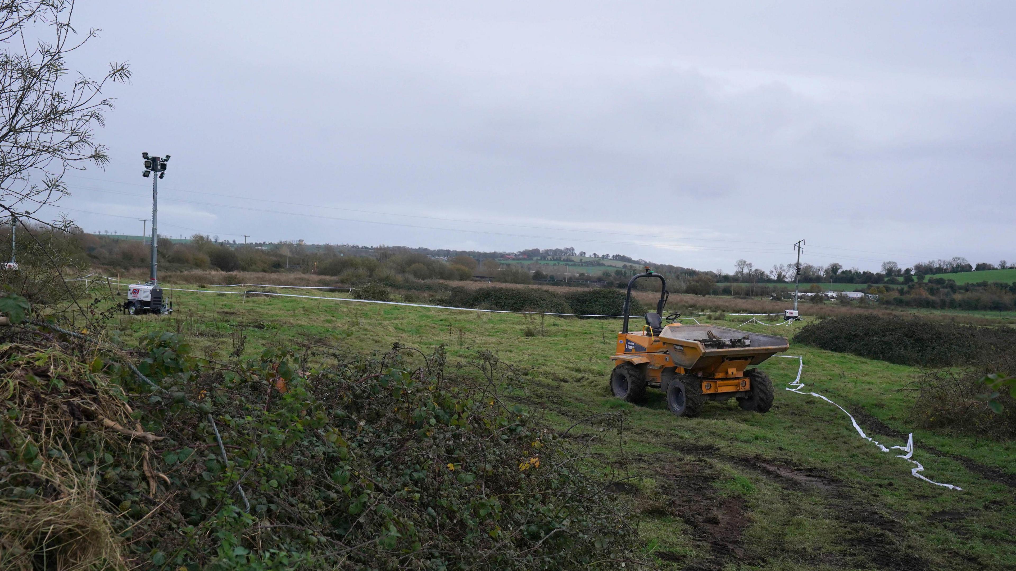 The image shows a countryside landscape of green fields and bushes in the foreground. Off to the right of the image is a yellow digger-type piece of machinery, to the left is a grey coloured flood light that is turned off. The sky is a slate grey colour. 