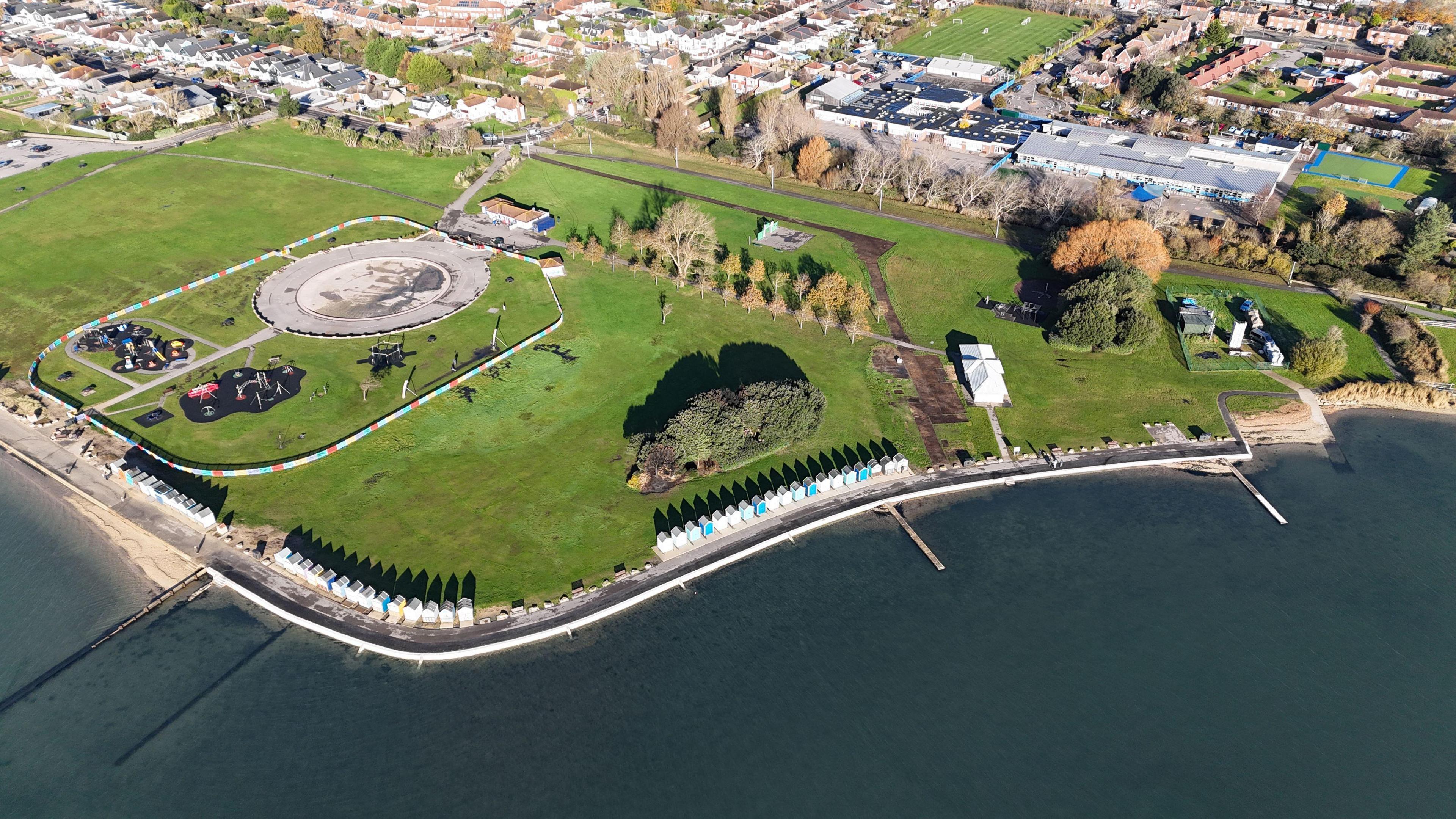 A drone photo of a large grassy park next to the sea. There's a road and a row of beach huts separating the park from the sea.