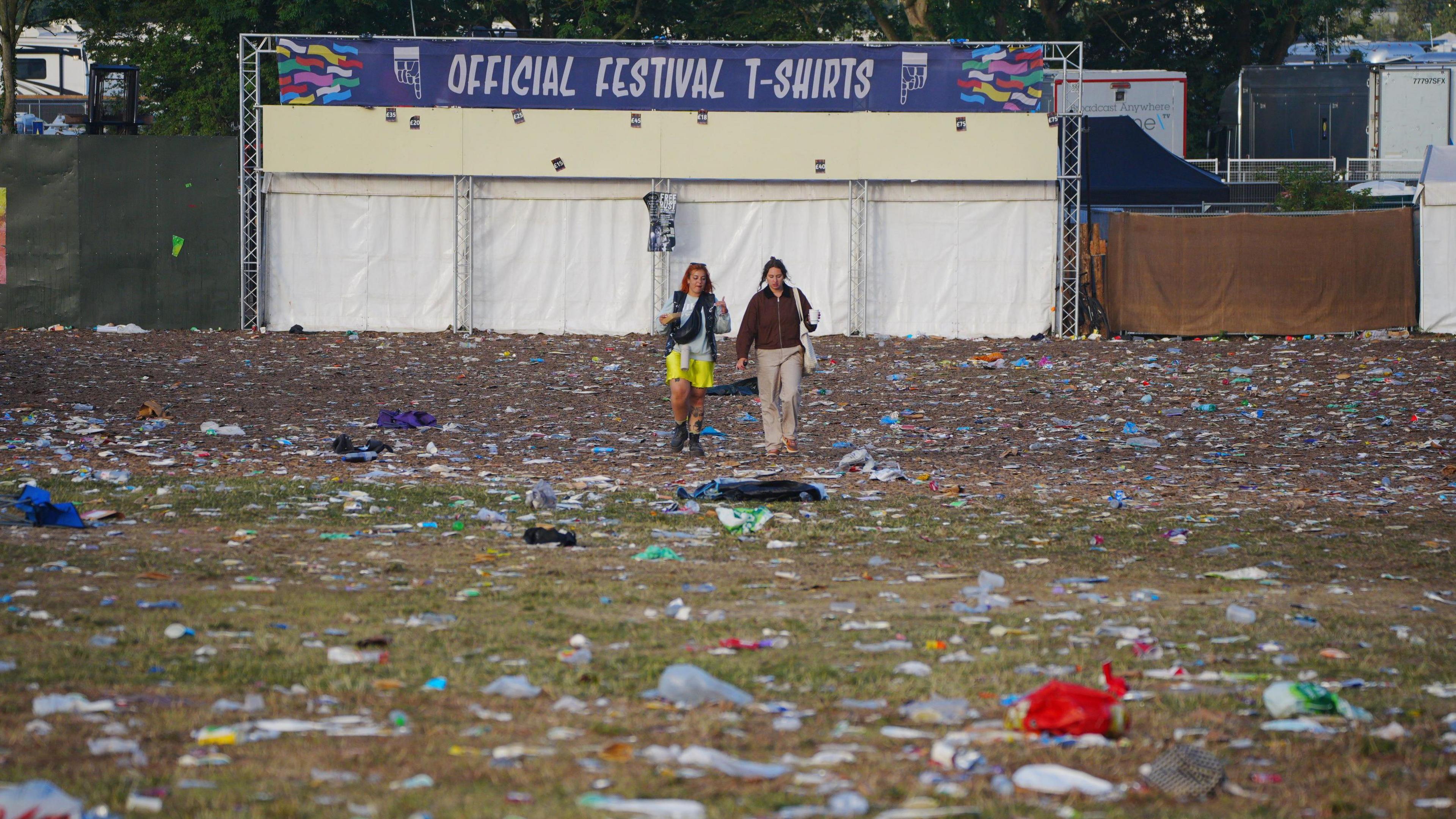 Two people stood in a field at Glastonbury Festival. They are both carrying bags. There is rubbish all over the ground. There is a T-shirt vendor which is closed up behind them.