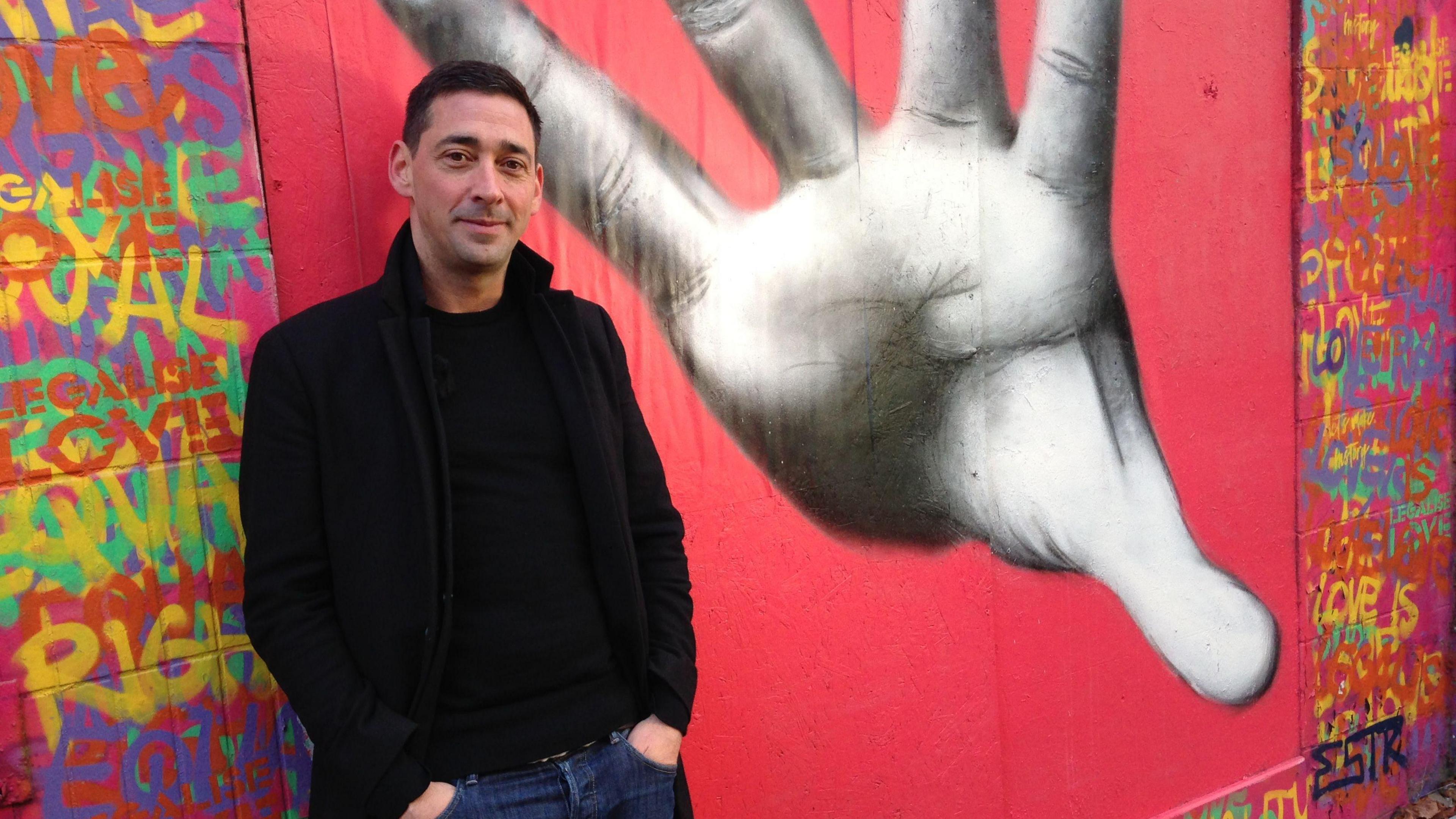 Colin Murray has black hair, is wearing a black jacket and black jumper. He is standing in front of a mural of a hand. 