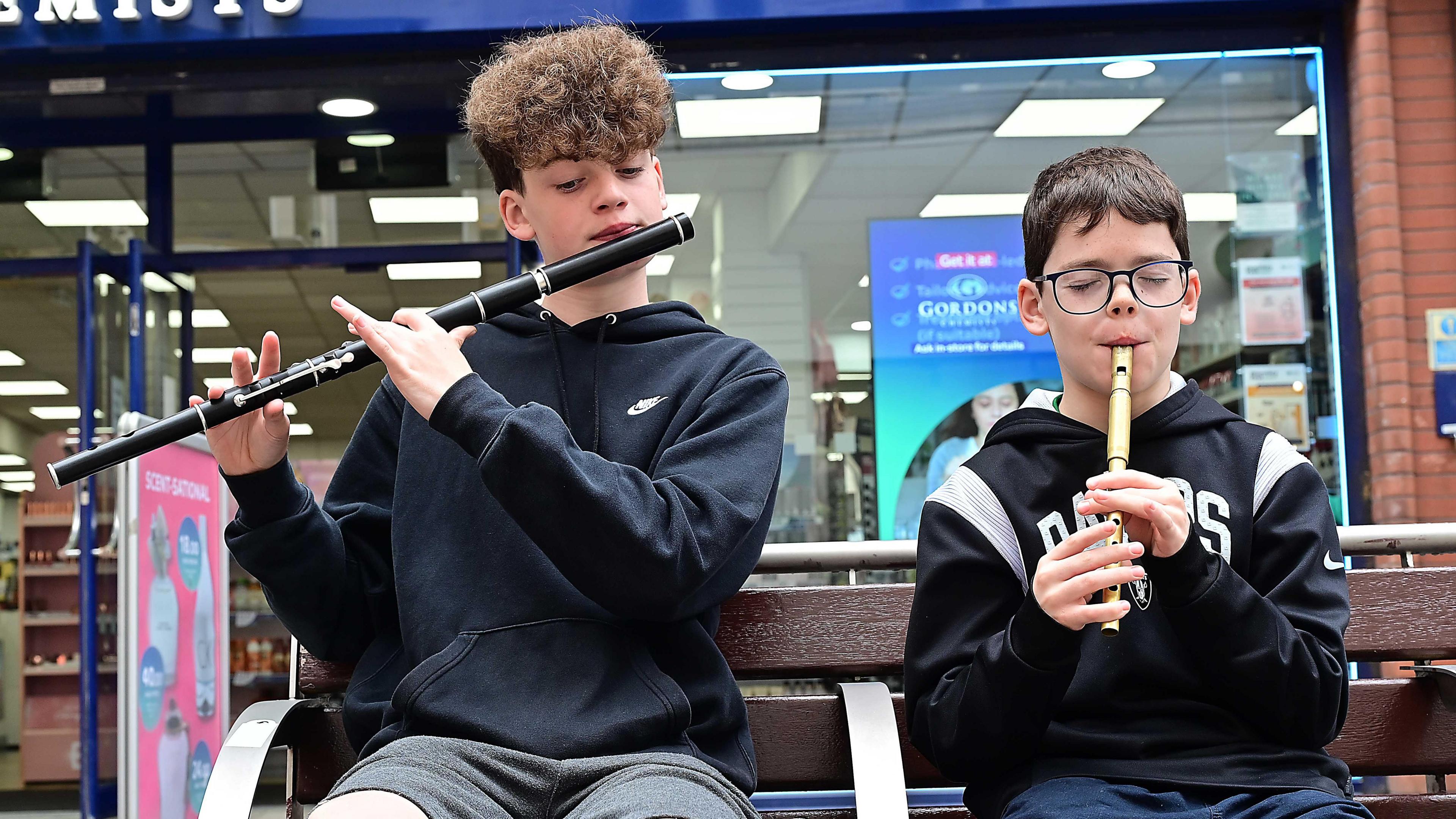 Two young boys with brown hair, wearing black hoodies, are playing wind instruments on a bench