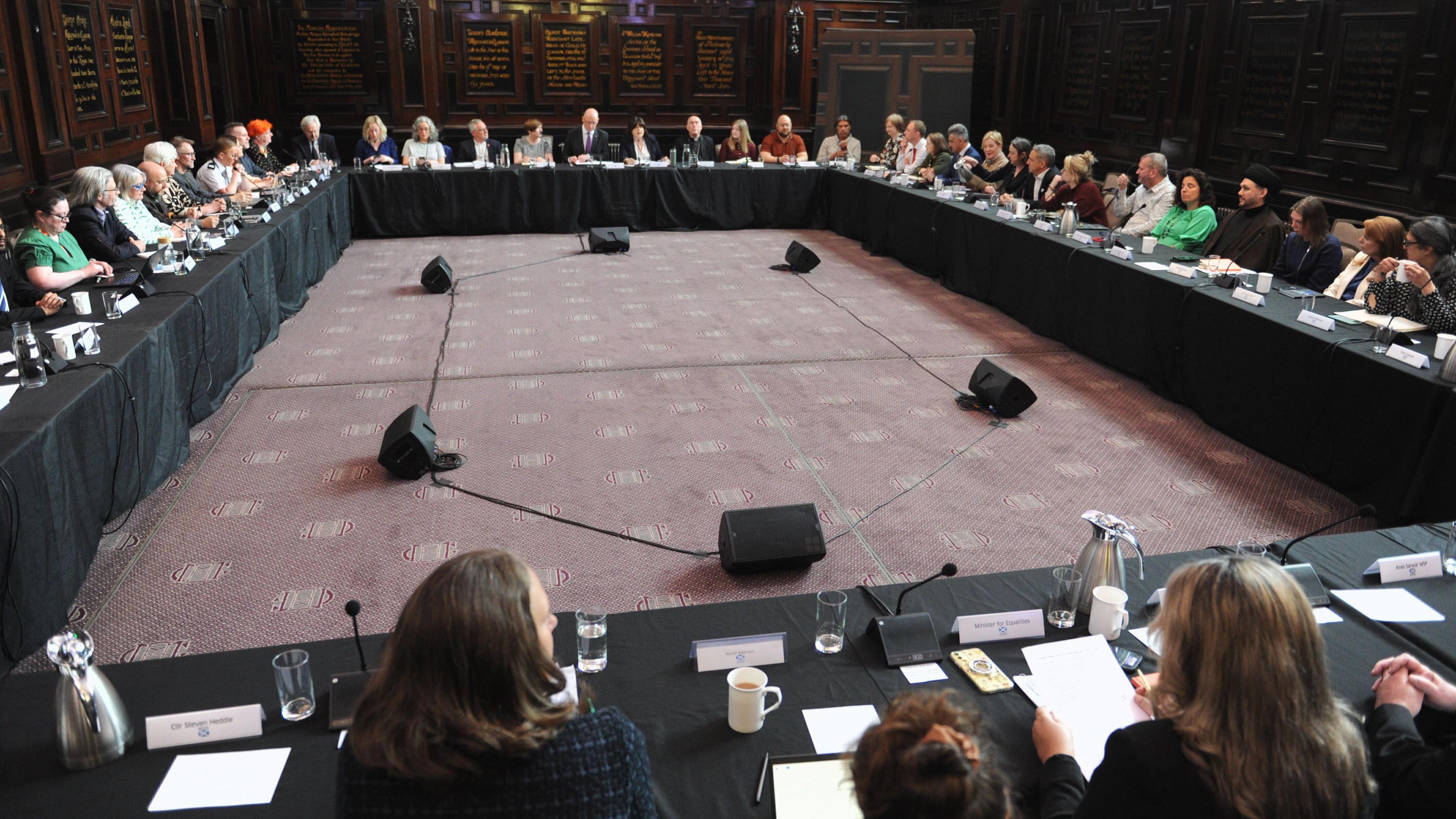 Four tables form a rectangle, with black drapes covering each one. Dozens of people are sitting behind them, with various microphones, papers and cups of coffee and glasses of water on the tables themselves.