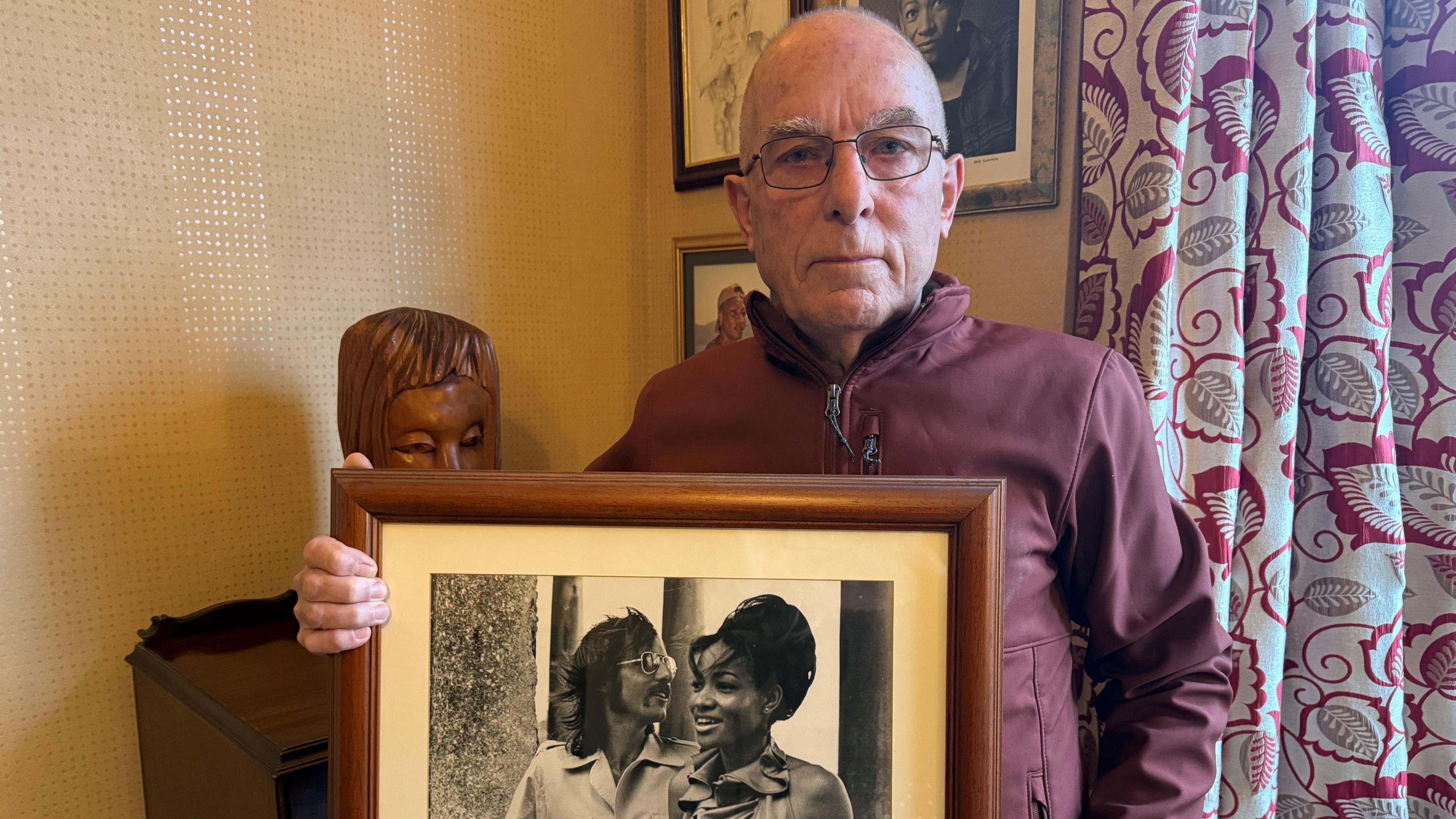 Dave Swaffer, a balding white man, stood in his living room wearing glasses holding an photo of him and his wife Judith.