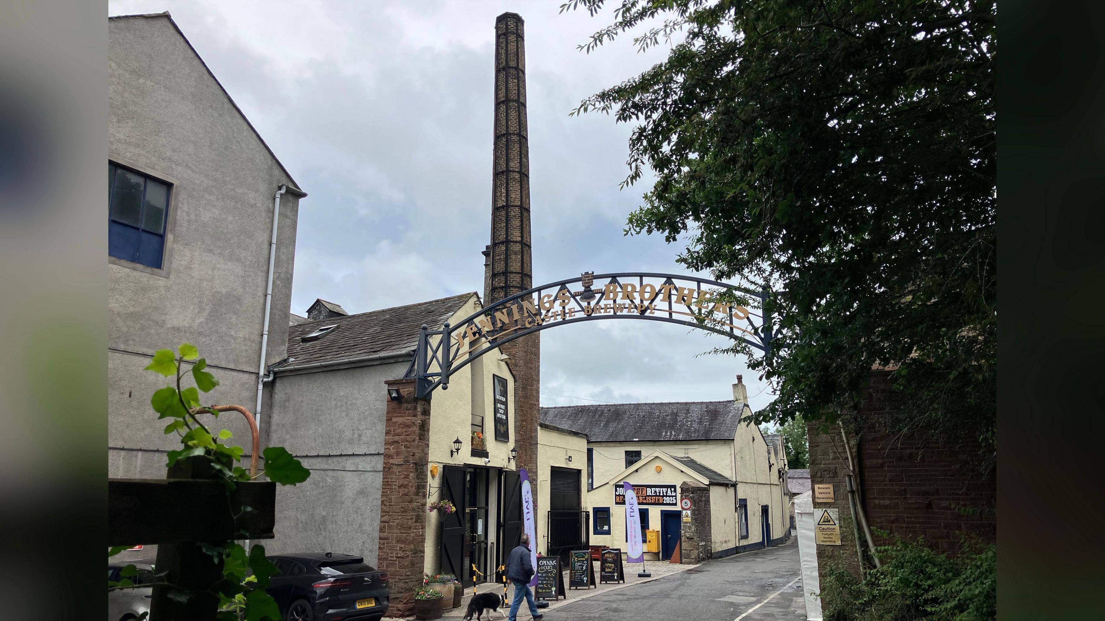 The exterior of Jennings Brewery. A tall brick chimney rises out of a two-storey yellow building, which has multiple signs on it. A metal arch over the entryway reads: "Jennings Brothers".