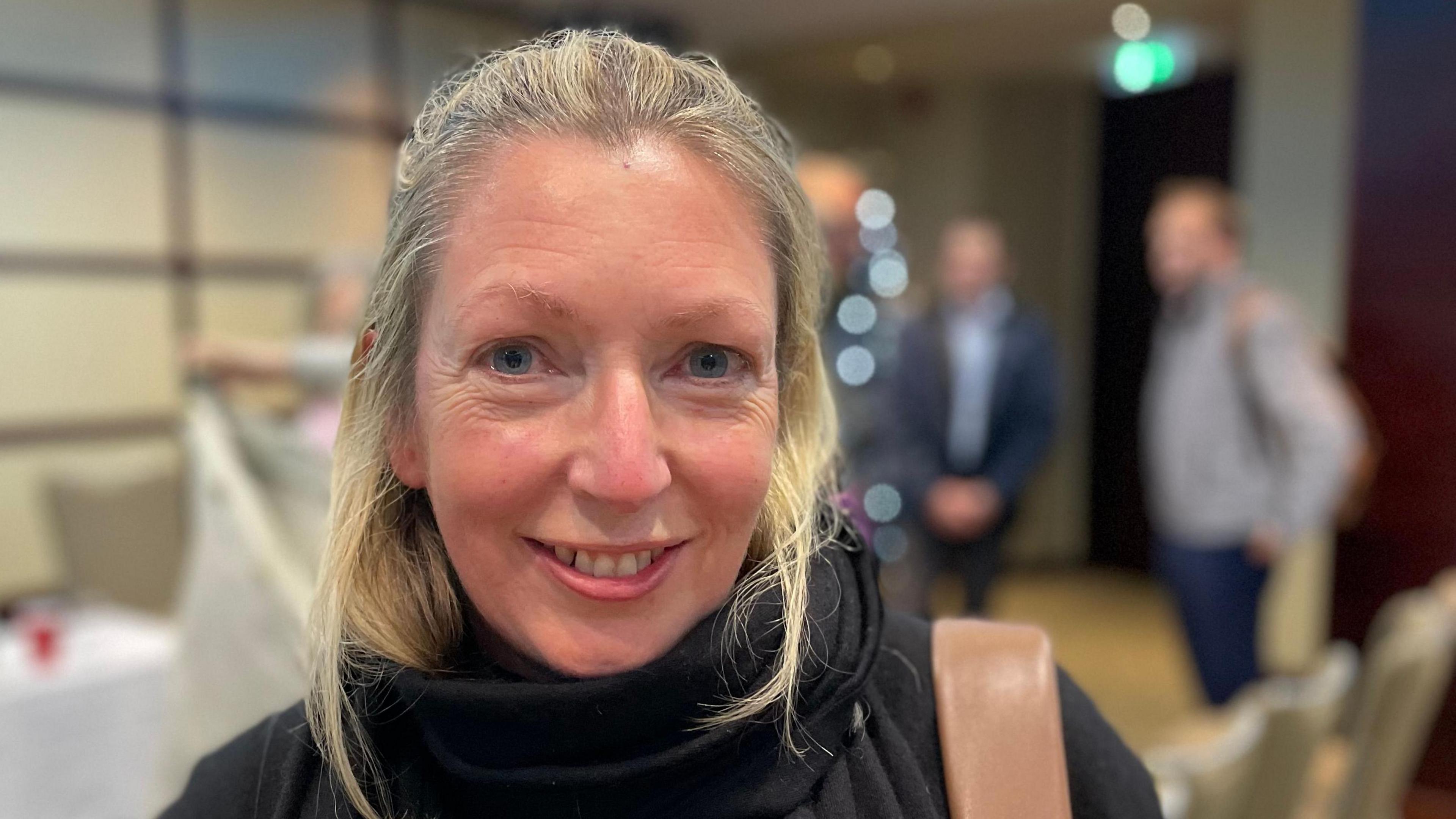 Alison Horton is a woman with blonde hair and blue eyes. She is smiling into the camera. She has a black top and scarf on and a brown back strap on her right shoulder, as you look at the photo. The background is an out-of-focus meeting room.