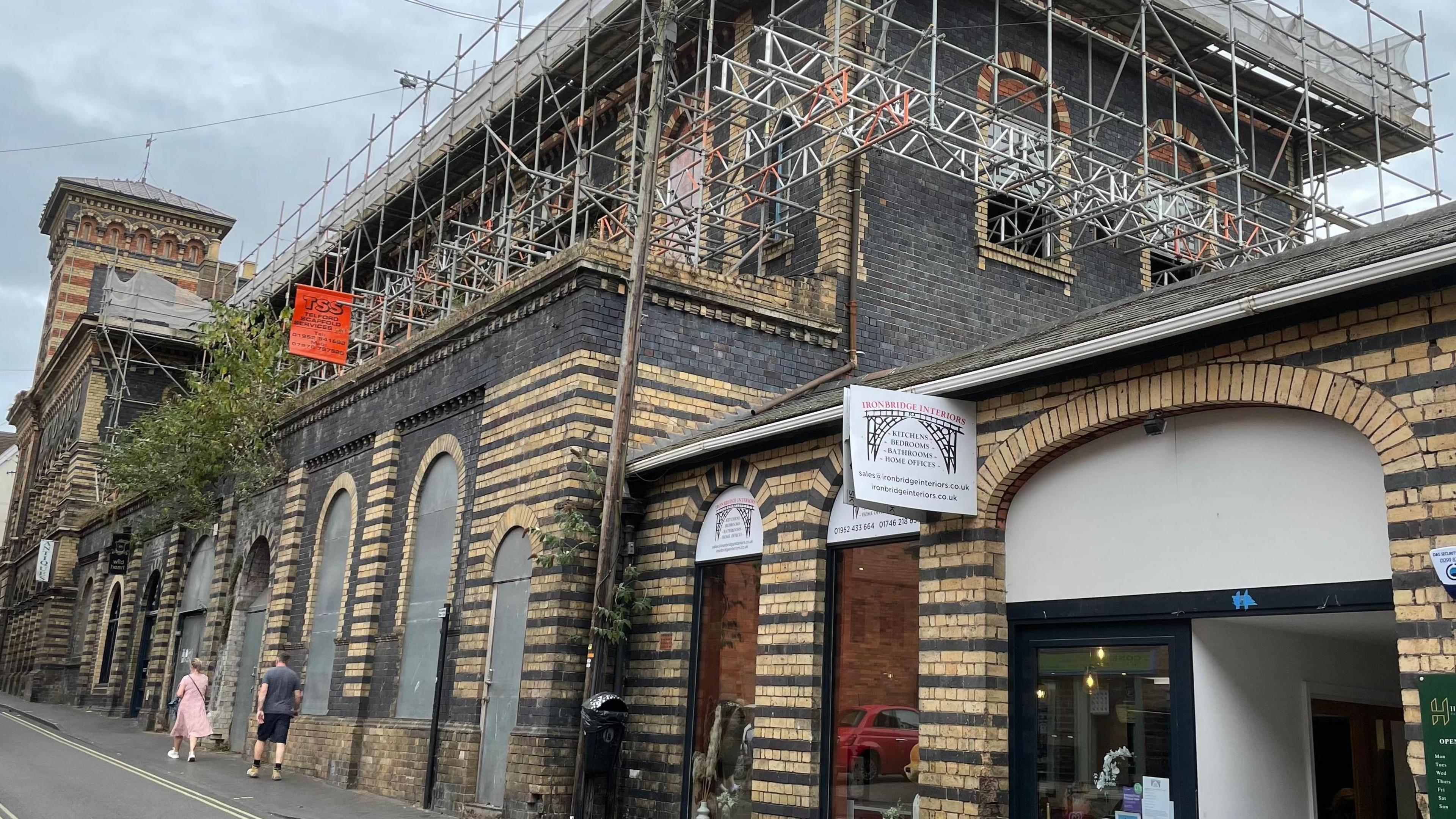 The New Market buildings, which are of Italianate architecture, with scaffolding on its upper floors and arches boarded up with metal sheets. People can be seen walking up the street past it. A tower at the far end is topped by a weather vane and another business is open next door.
