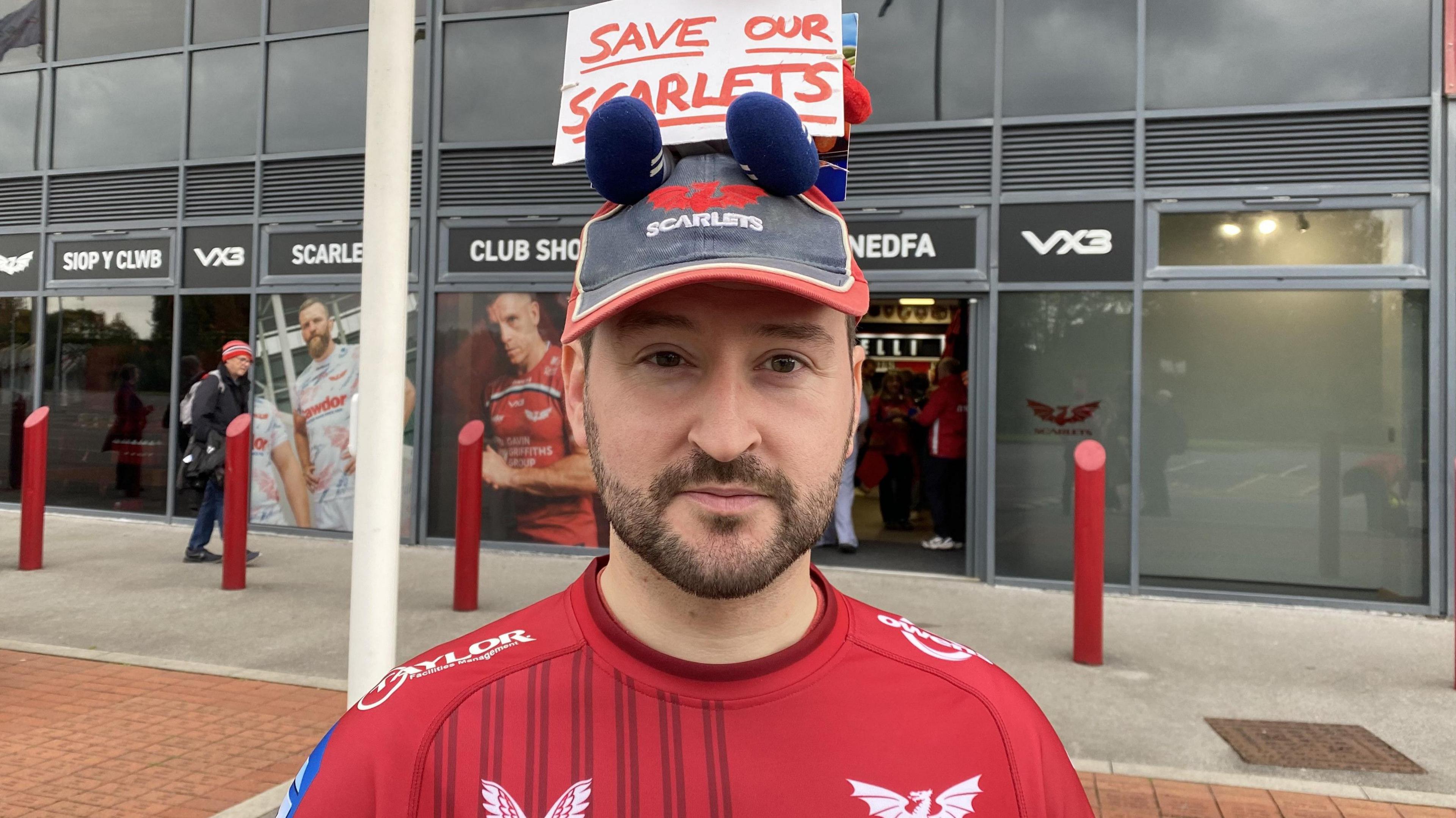 Tomos Davies is wearing a Scarlets Jersey and a Scarlets baseball cap. The cap has a sign which says 'Save our Scarlets' written in red capital letters on it. 