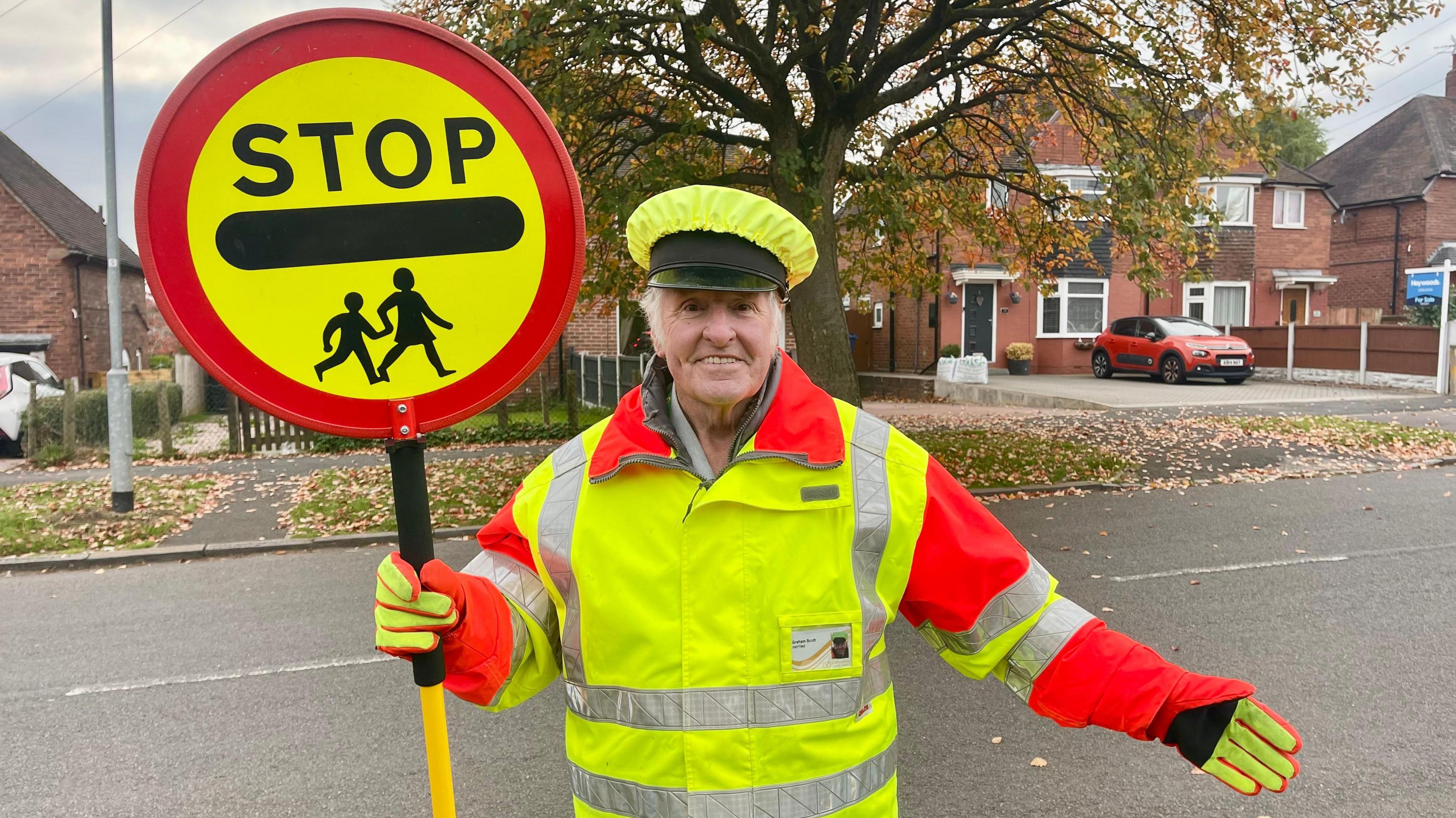 A man stands beside a road dressed in a high vis peaked hat, coat and gloves holding a STOP sign. He has white hair and is wearing a grey shirt beneath his uniform. On the other side of the road is a large tree, pavement, grassy areas, semi-detached houses and a red car on a driveway.