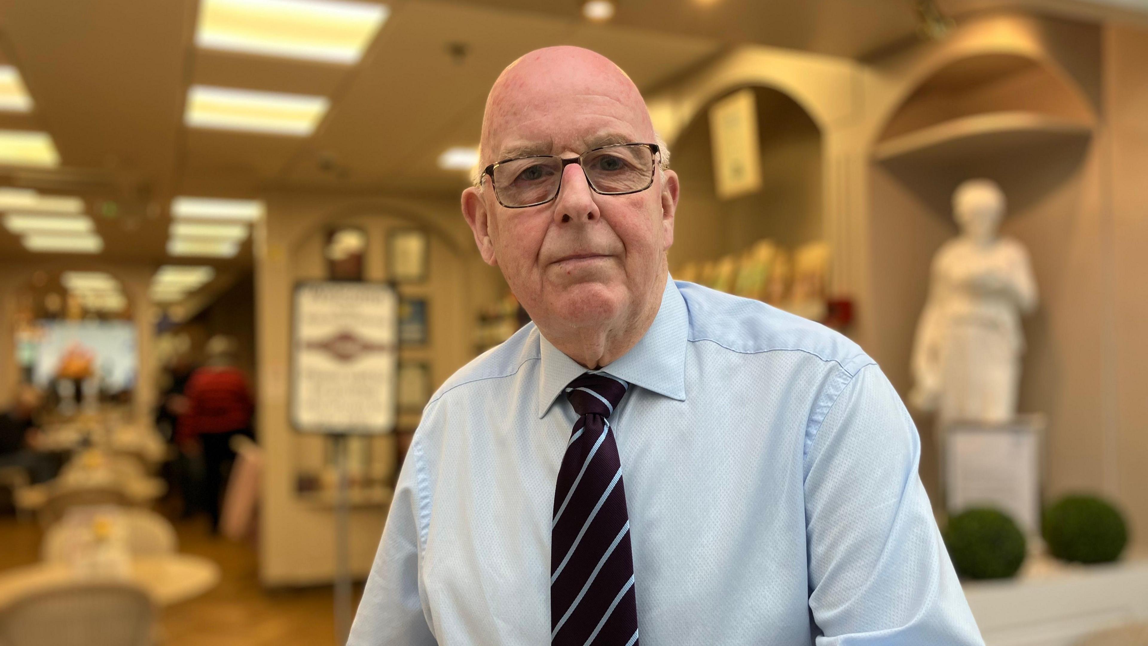 Tony Cunningham is outside his tea room. Bald and with glasses, he is wearing a light blue shirt and a brown-and-blue striped tie. 