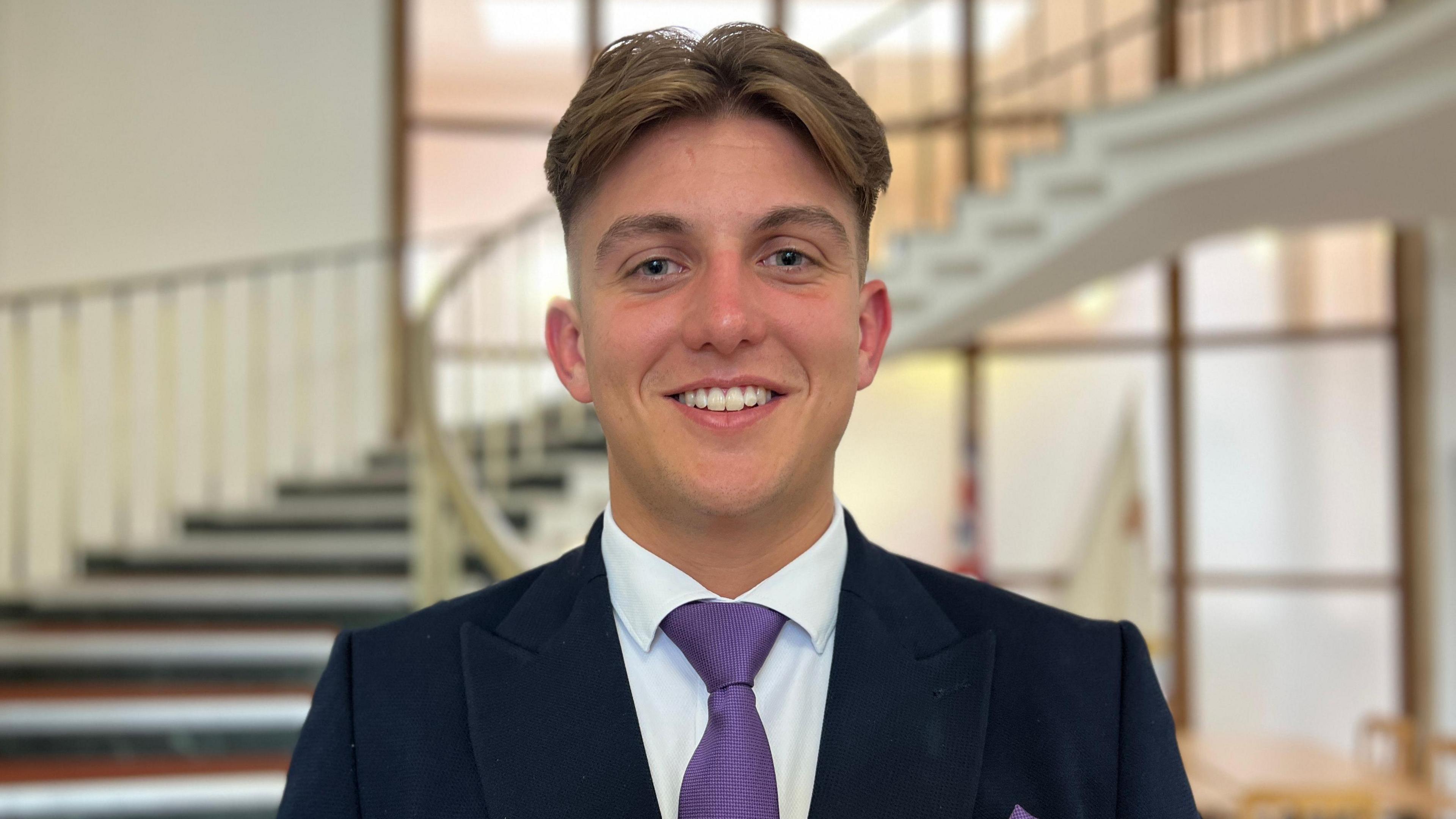 George Finch is pictured in front of a spiral staircase. He is wearing a navy suit, white shirt and purple tie. He has light brown hair brushed into a centre parting.