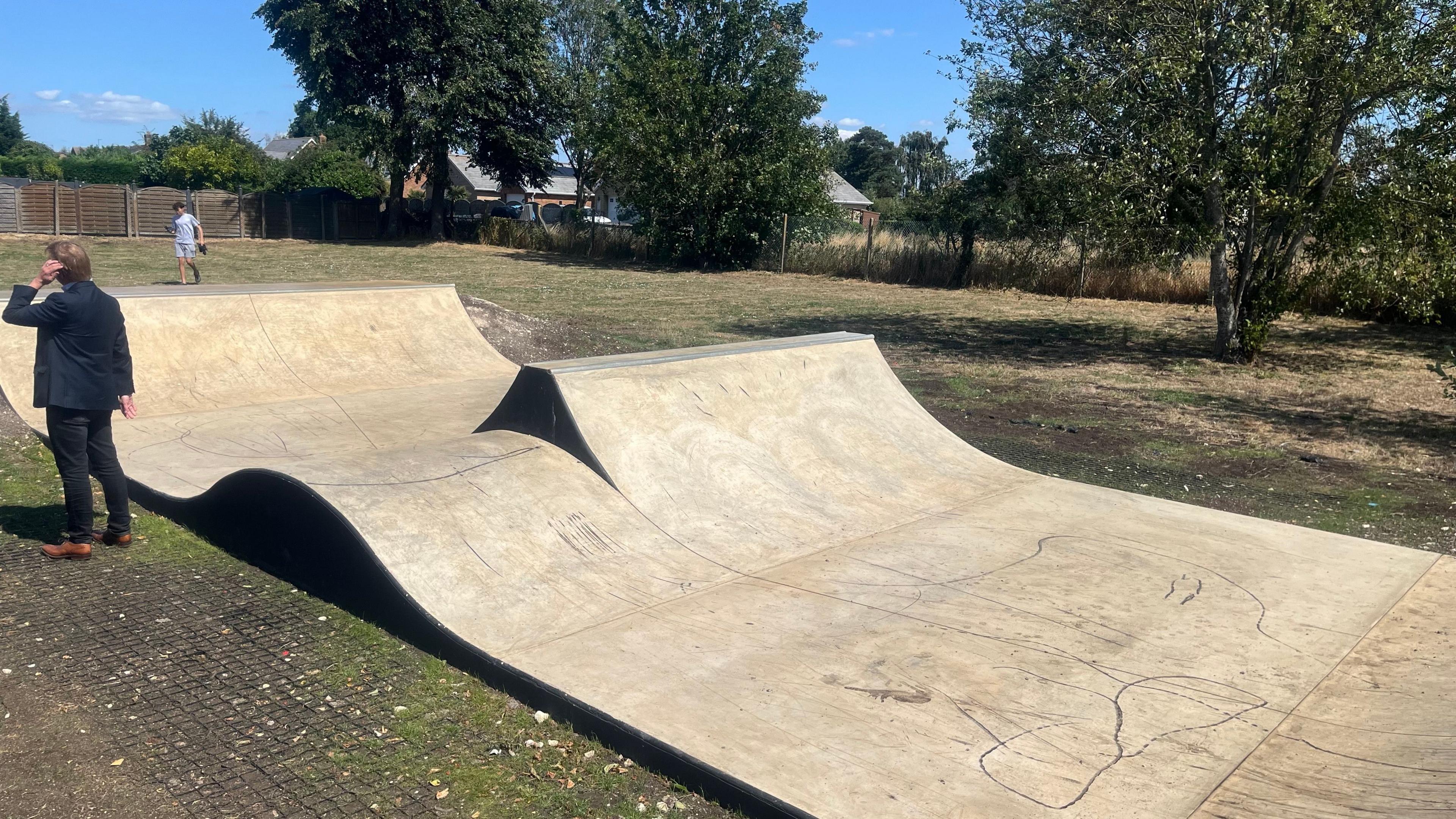 The new skate ramp is a long wooden platform with a small hill in the centre on the left side. On the fight is a steeper hill with a narrow flat platform at the top. It is situated in a grassy area with trees. 