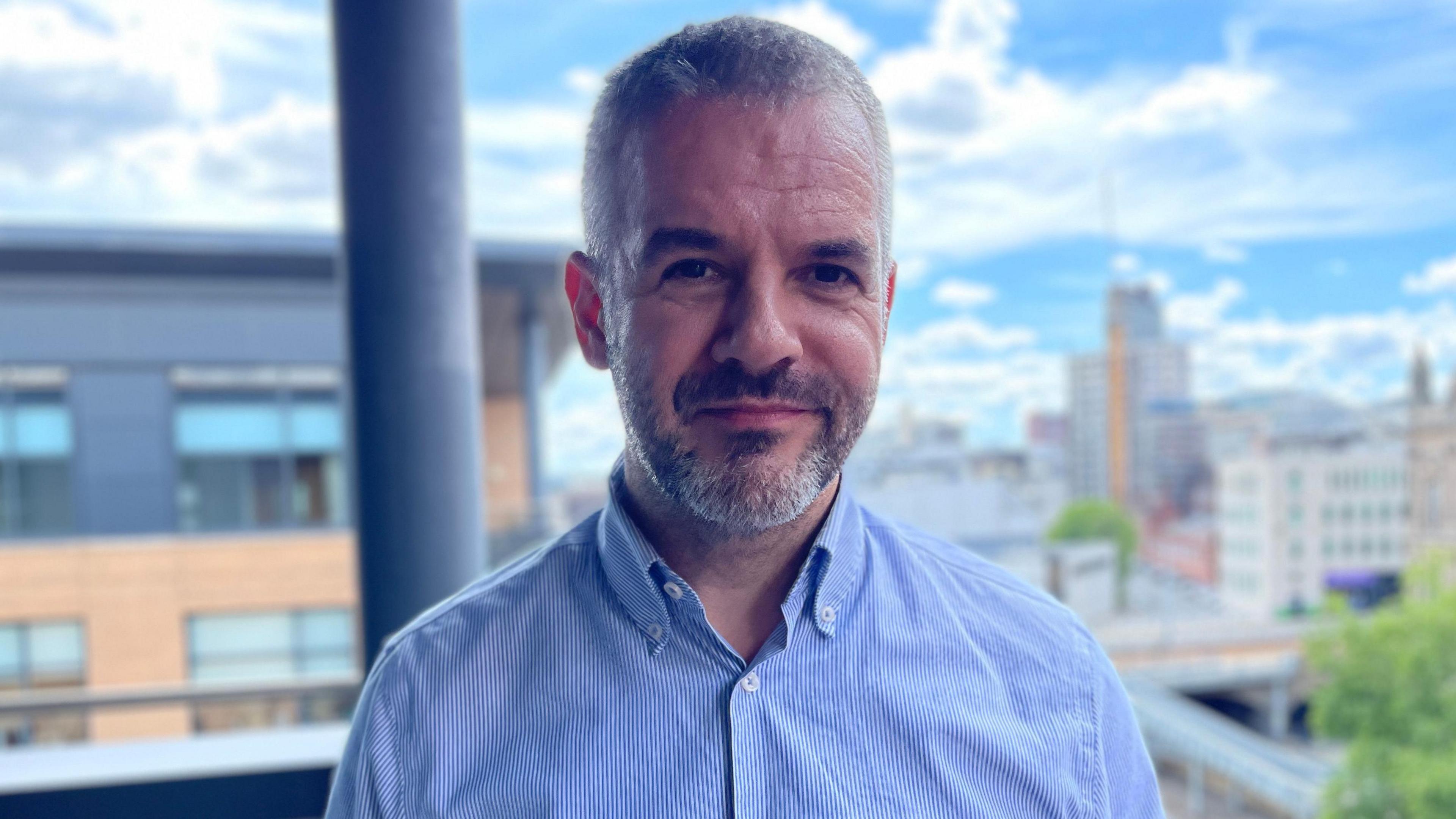 A man with short grey-black hair and a short beard, wearing a blue shirt.