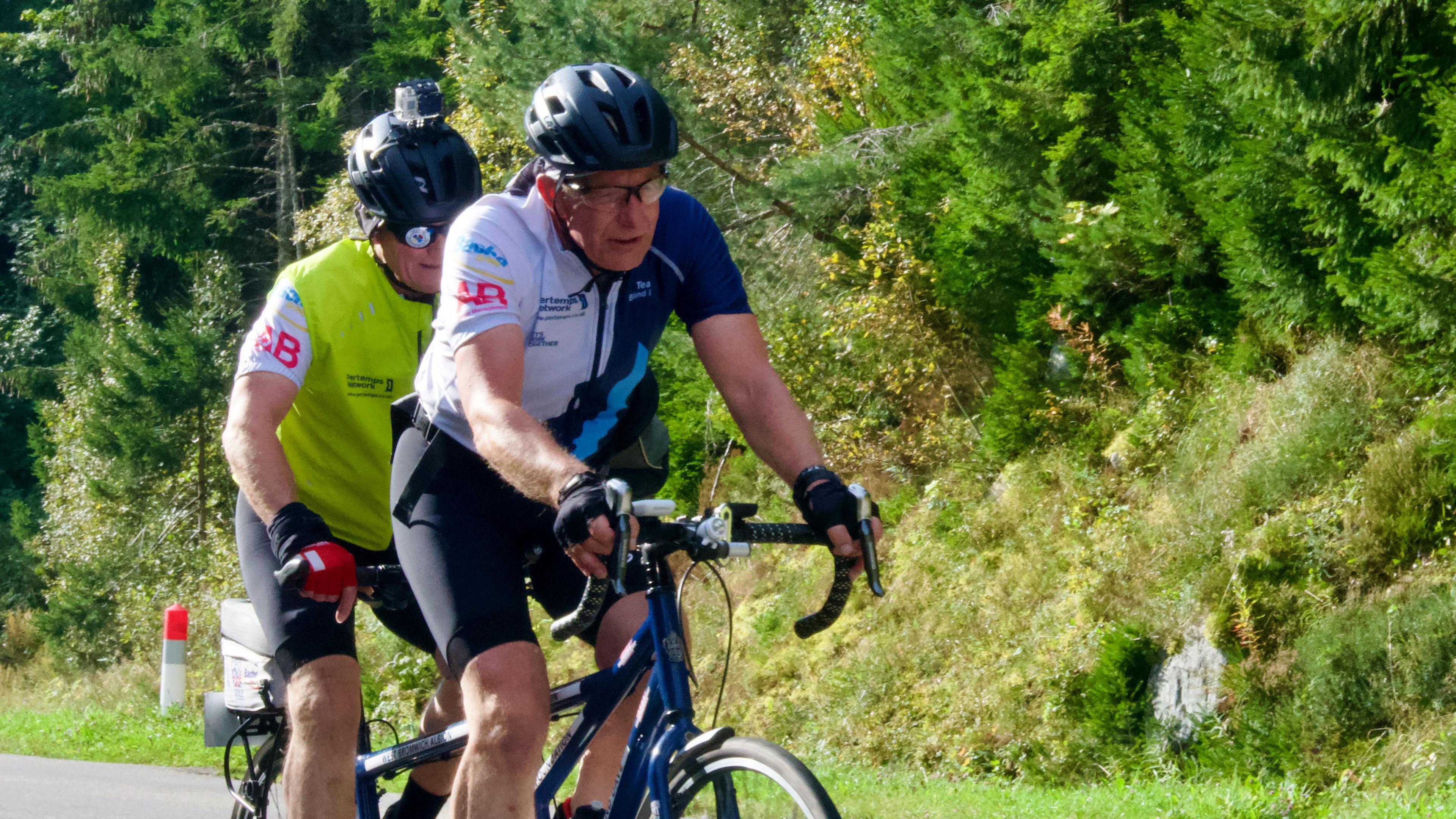 Two men on a tandem cycle. The front rider is wearing a blue and white halved cycling shirt and black cycle helmet. The back rider is a lime green gillet and a helmet with a camera on top. They are pedalling uphill with green woodland to their left