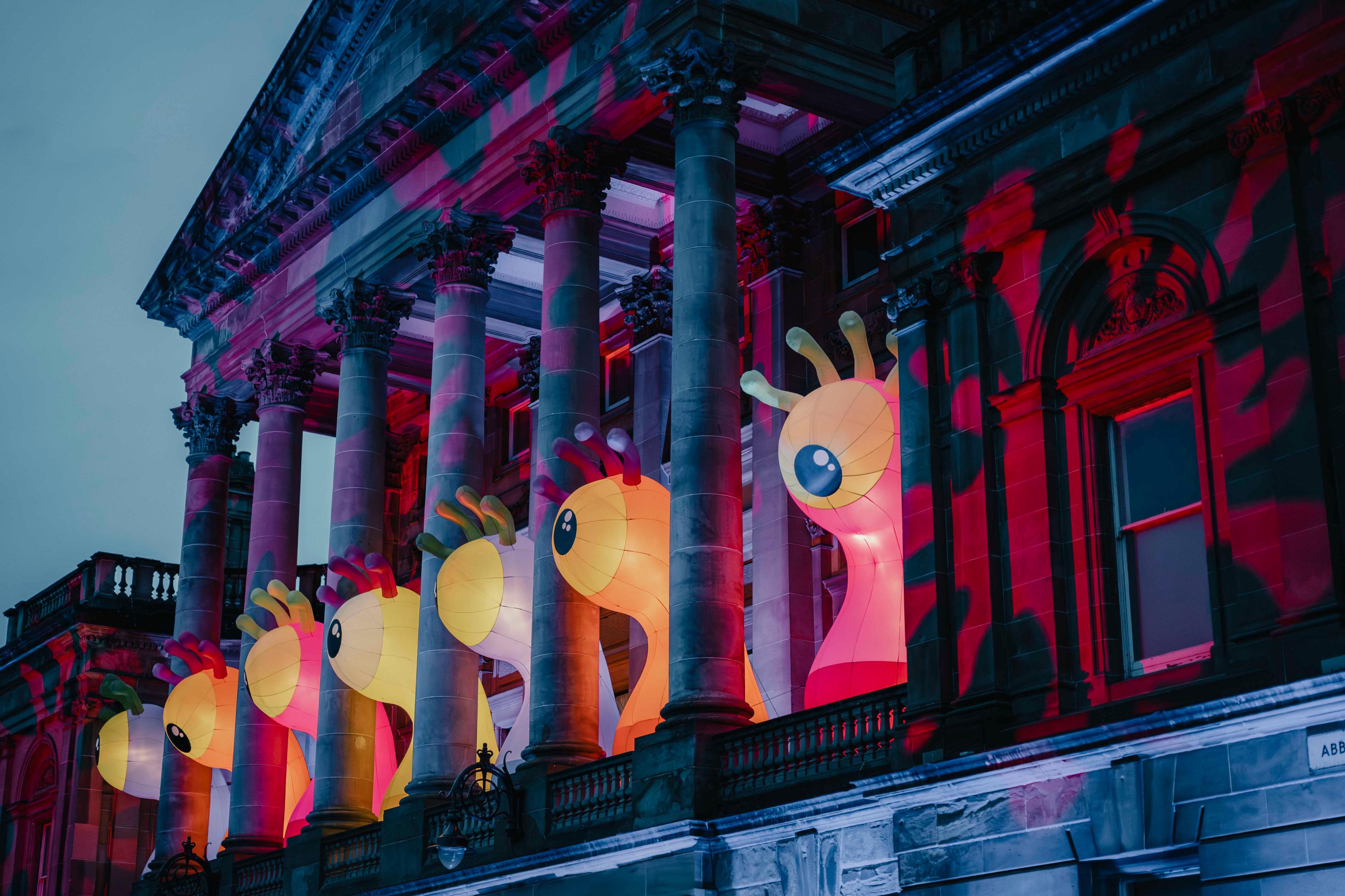 A mid 19th Century sandstone town hall building with pillars lit up by a purple and red light installation. Between the pillars are large inflatable eyeballs.