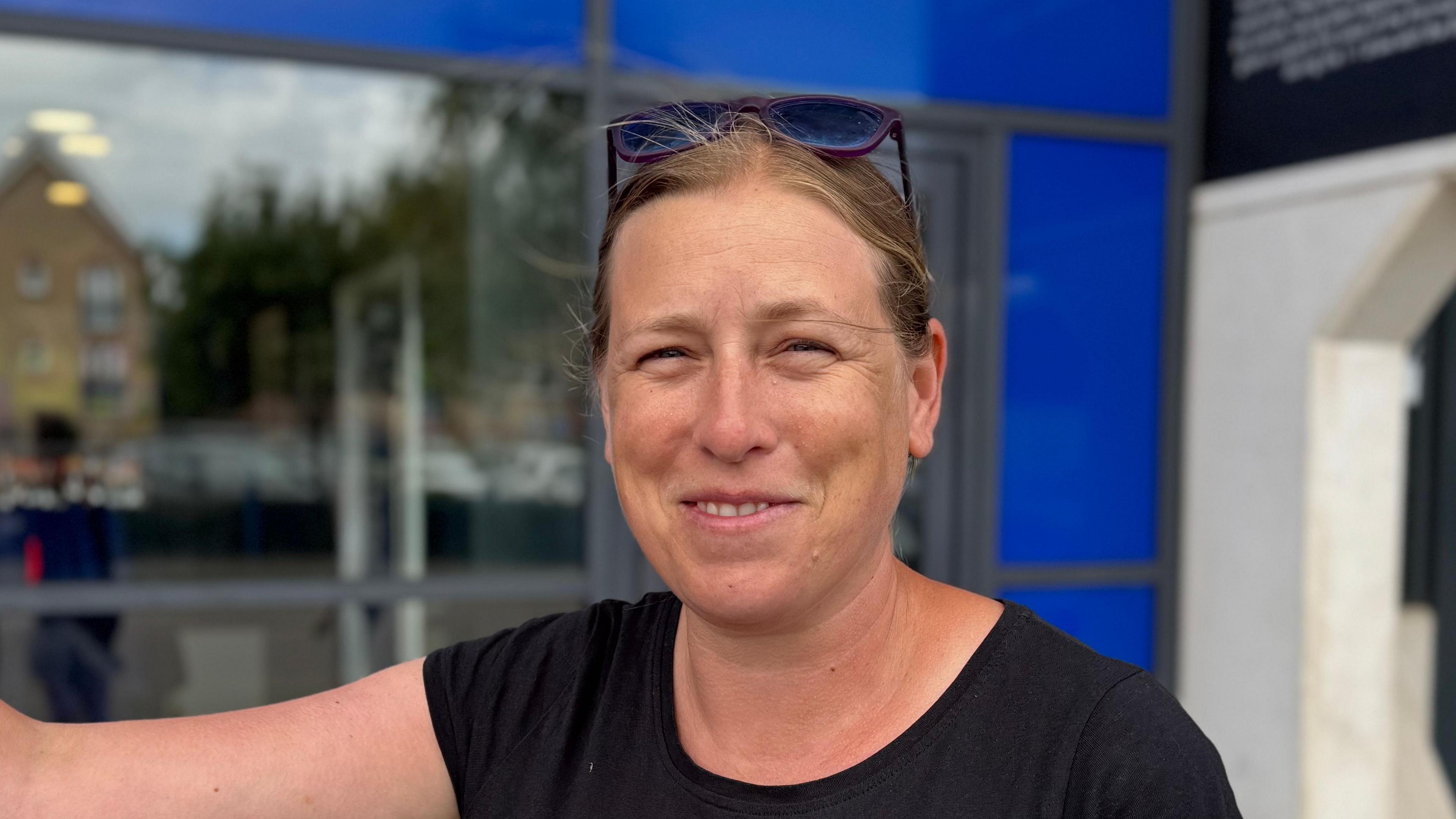 A head and shoulders picture of a woman. She is wearing a black top and her sunglasses are on her head. She is smiling into the camera.