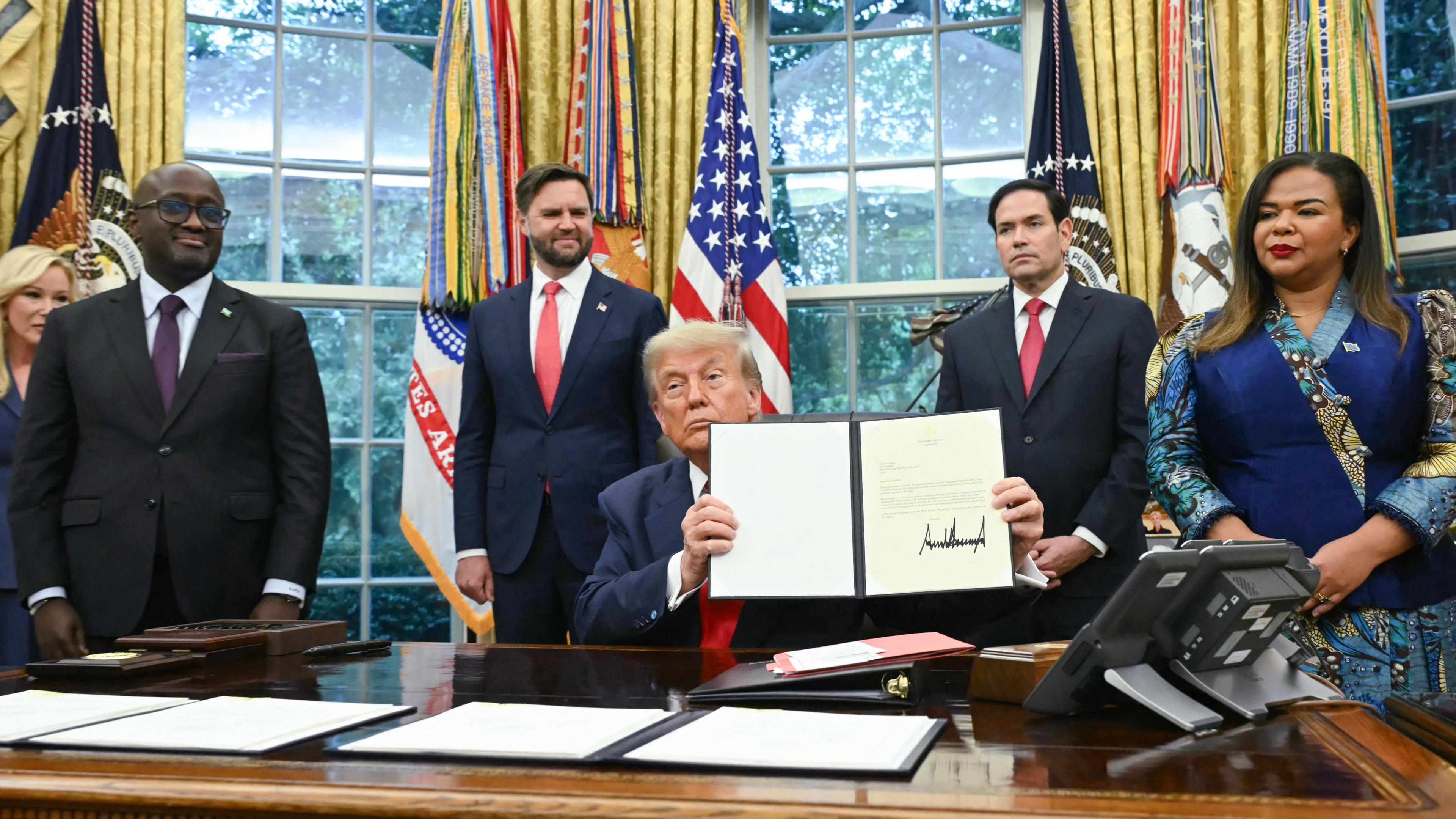 Donald Trump sits at a table in the Oval office holding up a signed letter. Standing next to him are officials from Rwanda and the Democratic Republic of Congo