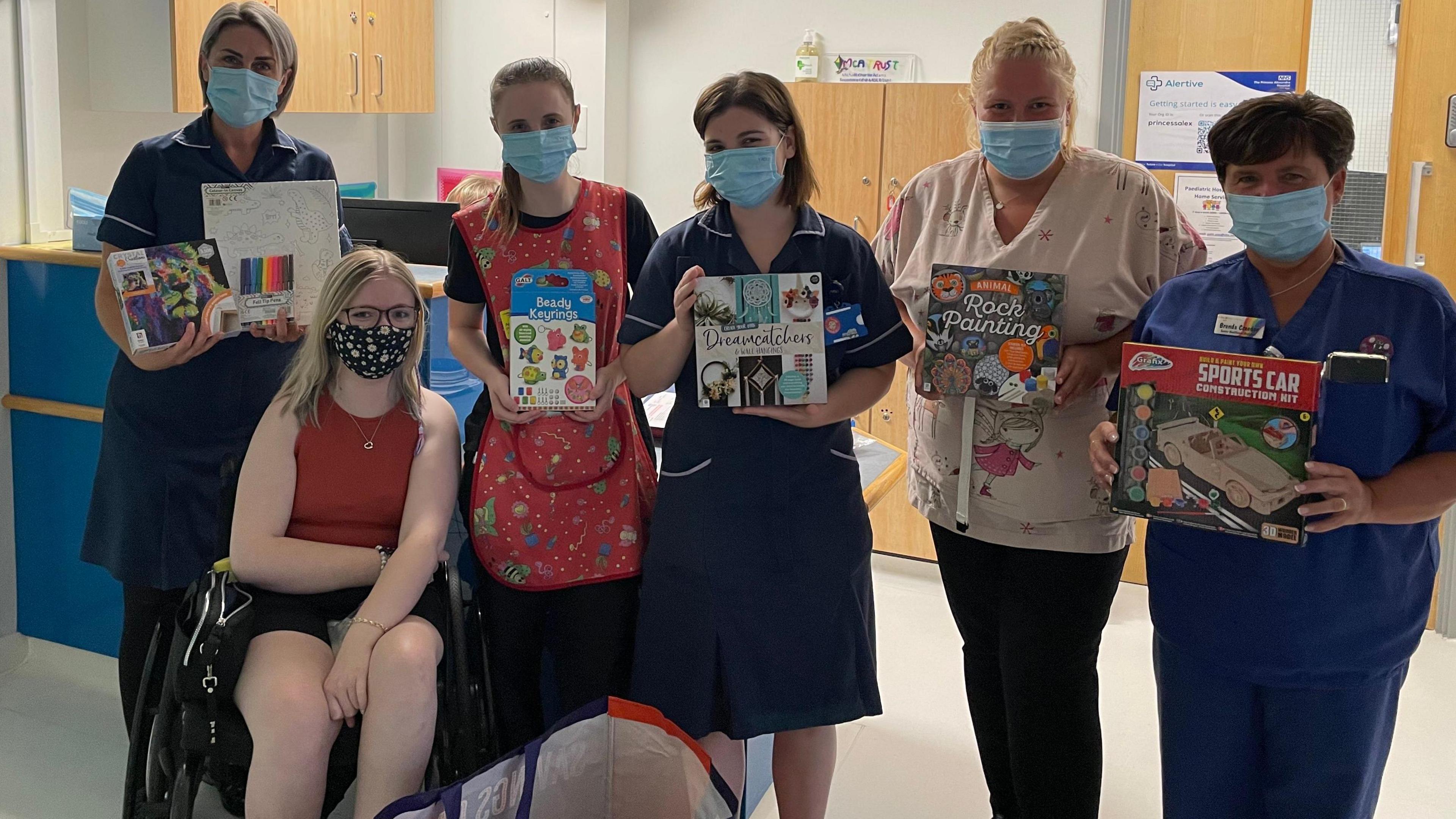 A group of nurses are holding books and art supplies in a hospital. Katie is sat nearby. Everybody is wearing a face mask.