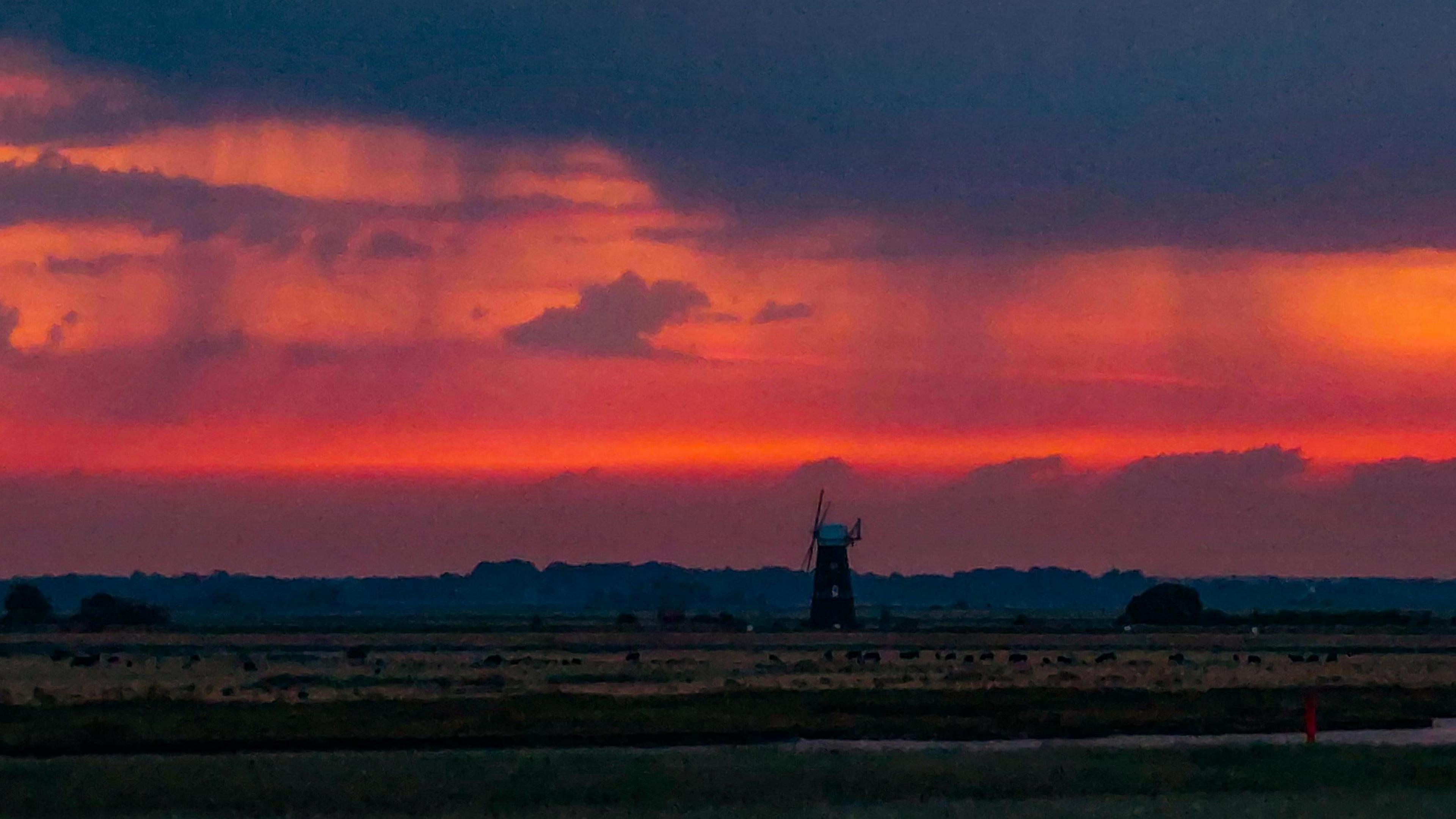 View out towards a windmill in the distance with cloudy skies and streaks of what looks like thin cloud beneath which is rain evaporating.  The setting sun providing the sky with a red backdrop.