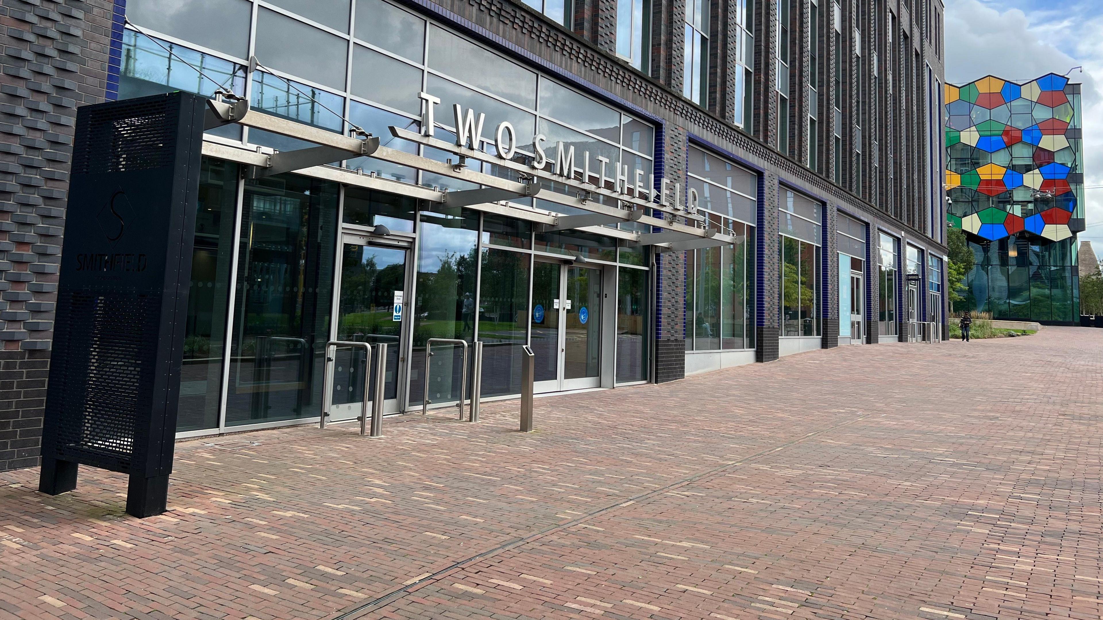 A modern office block in dark grey brick with extensive glazing. Cut-out metal letters above the entrance read "Two Smithfield".