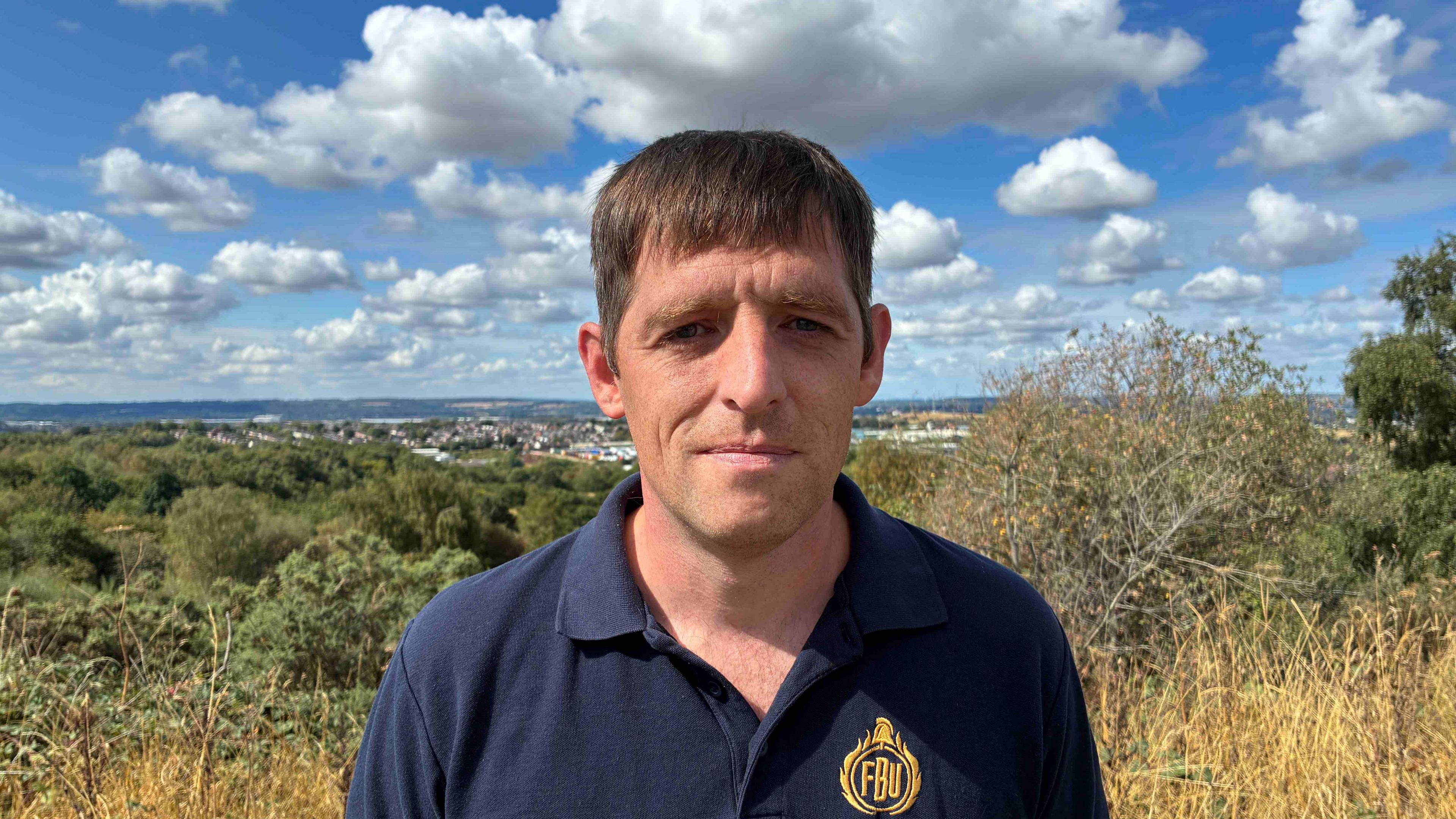 A close-up of a man with brown hair looking at the camera with a solemn expression, wearing a blue polo shirt that says FBU on it in gold thread. He stands in front of shrubbery and trees.