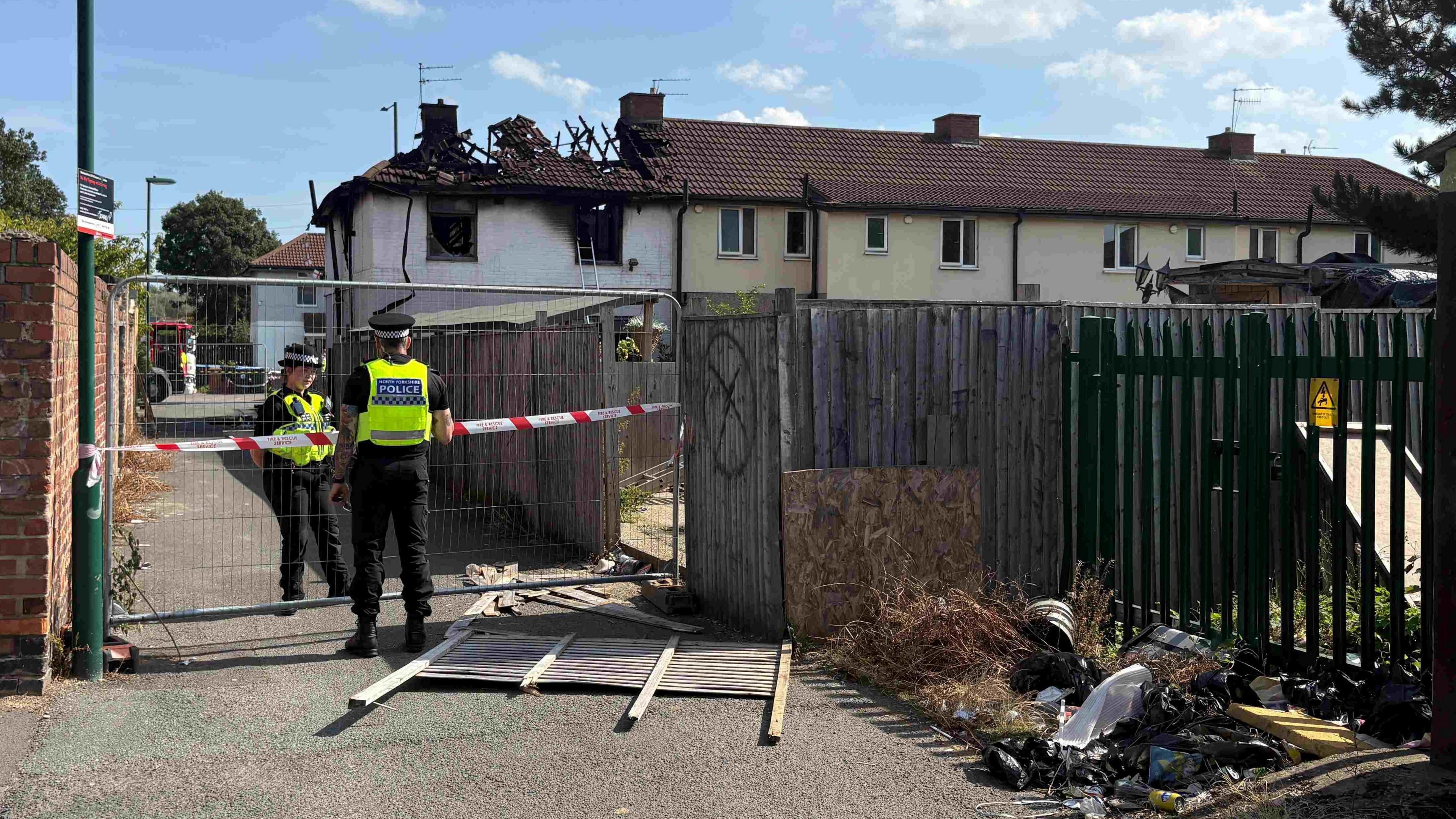 Police officers are standing at a metal fence sealing off an alleyway next to a badly burnt property. An 'x' in a circle can be seen next to a panel of fencing which is on the ground. The roof of the property is badly burnt.
