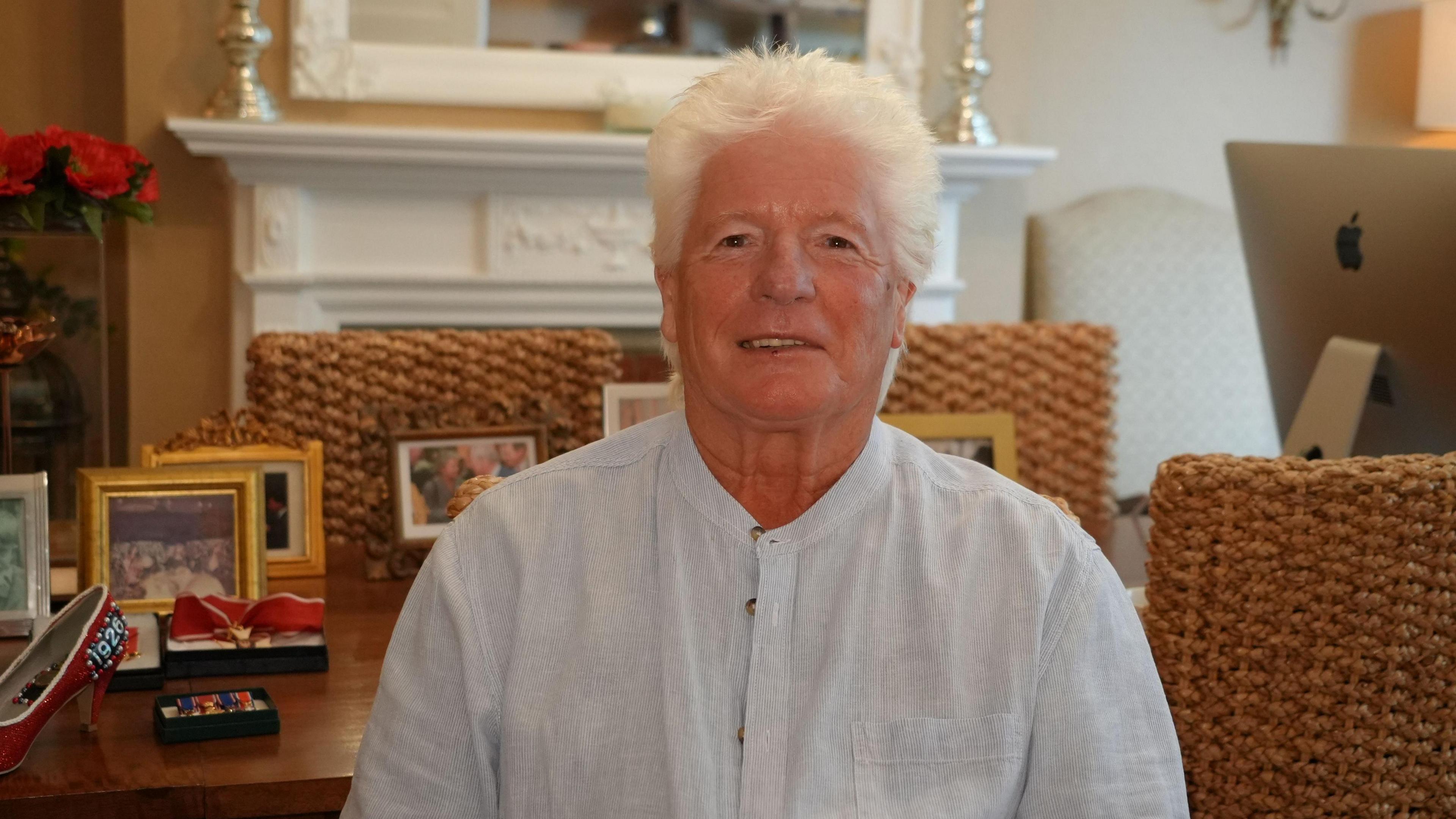 A man with thick white hair is seated on a wicker chair and smiles. He is wearing a very pale blue collarless shirt. Behind him, there are more wicker chairs, a large ornate white mantlepiece, a plant with a red flower and photographs on a large table.