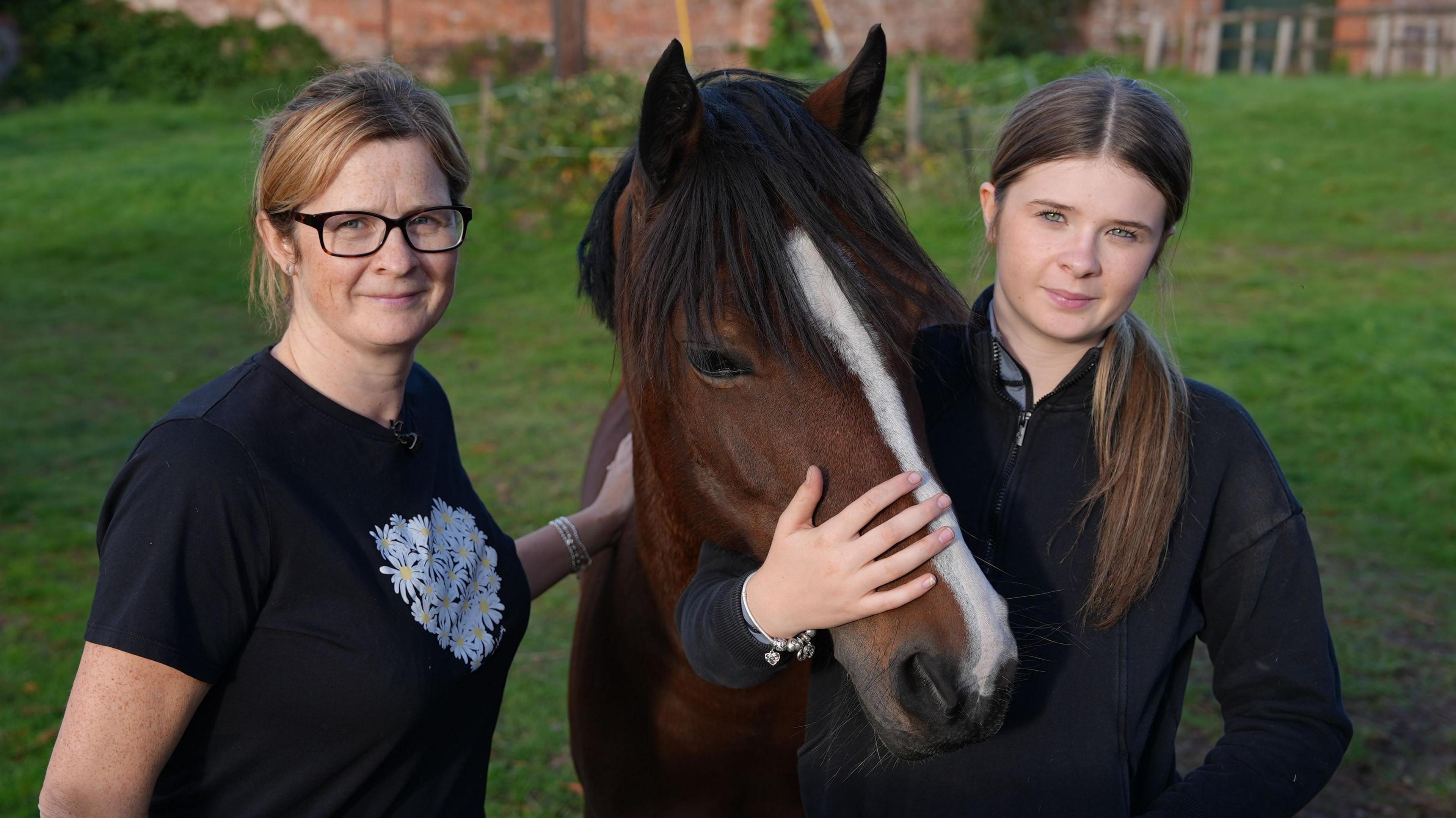 Alexandra Jones and her daughter Charlotte stand beside a dark brown horse with a white stripe down its nose. Charlotte gently rests a hand on the horse’s face and Alexandra strokes its back. The are stood in a grassy area with a red brick wall in the distance behind them that is out of focus. 