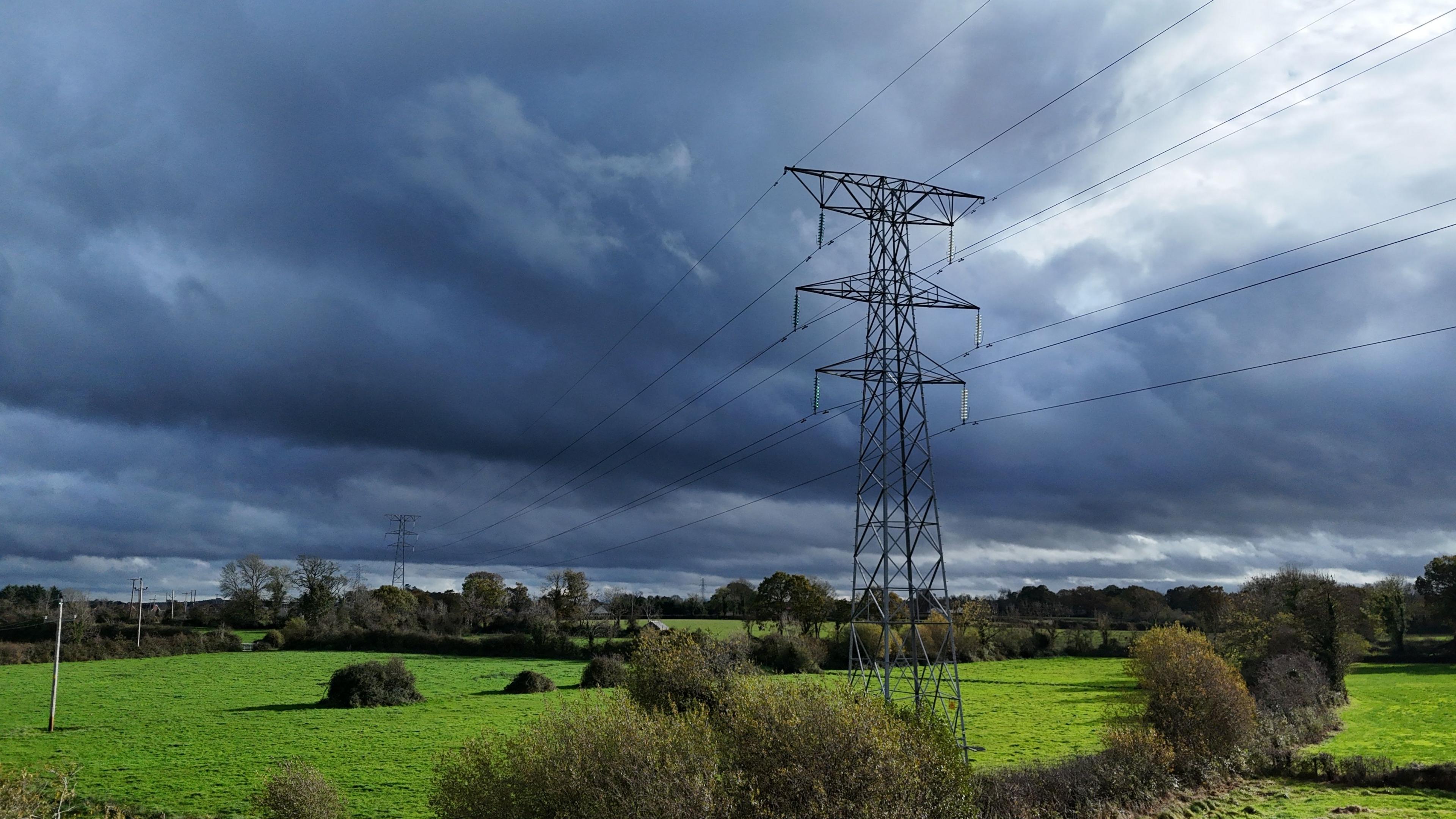 A pylon carrying power cables stands in a green field surrounded by flat fields, with hedges and small trees running between them. In the background, the sky is filled with large dark grey clouds. 