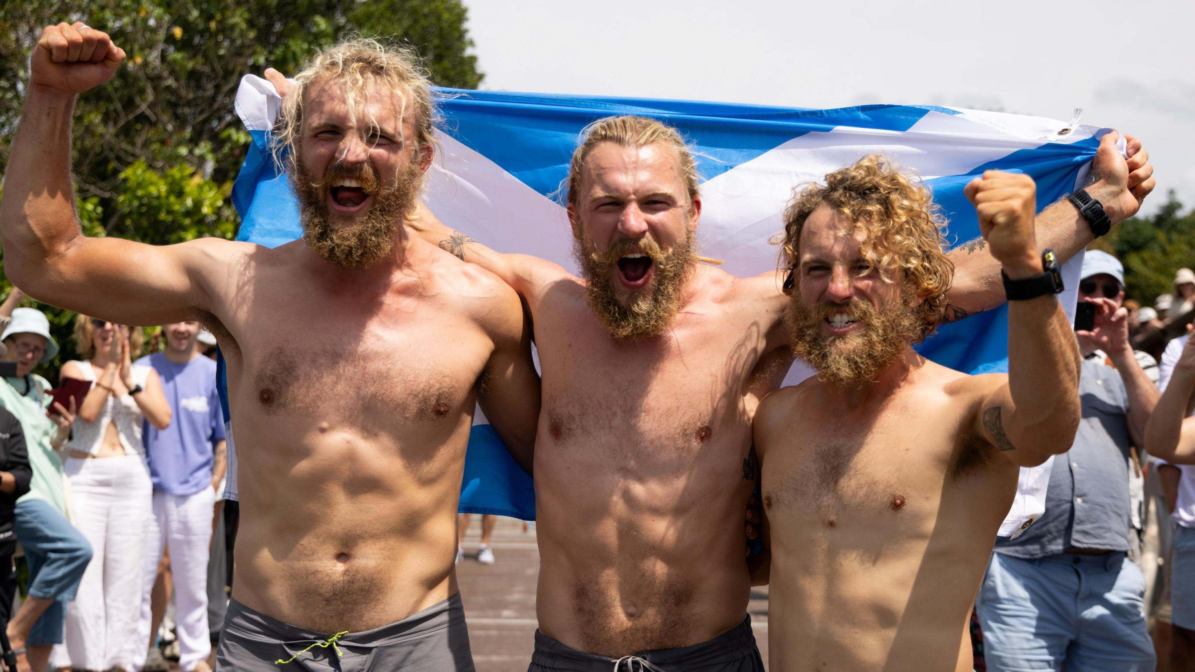 Three happy, bearded, barechested men stand in the sunshine in Cairns and wave their fists in celebration.