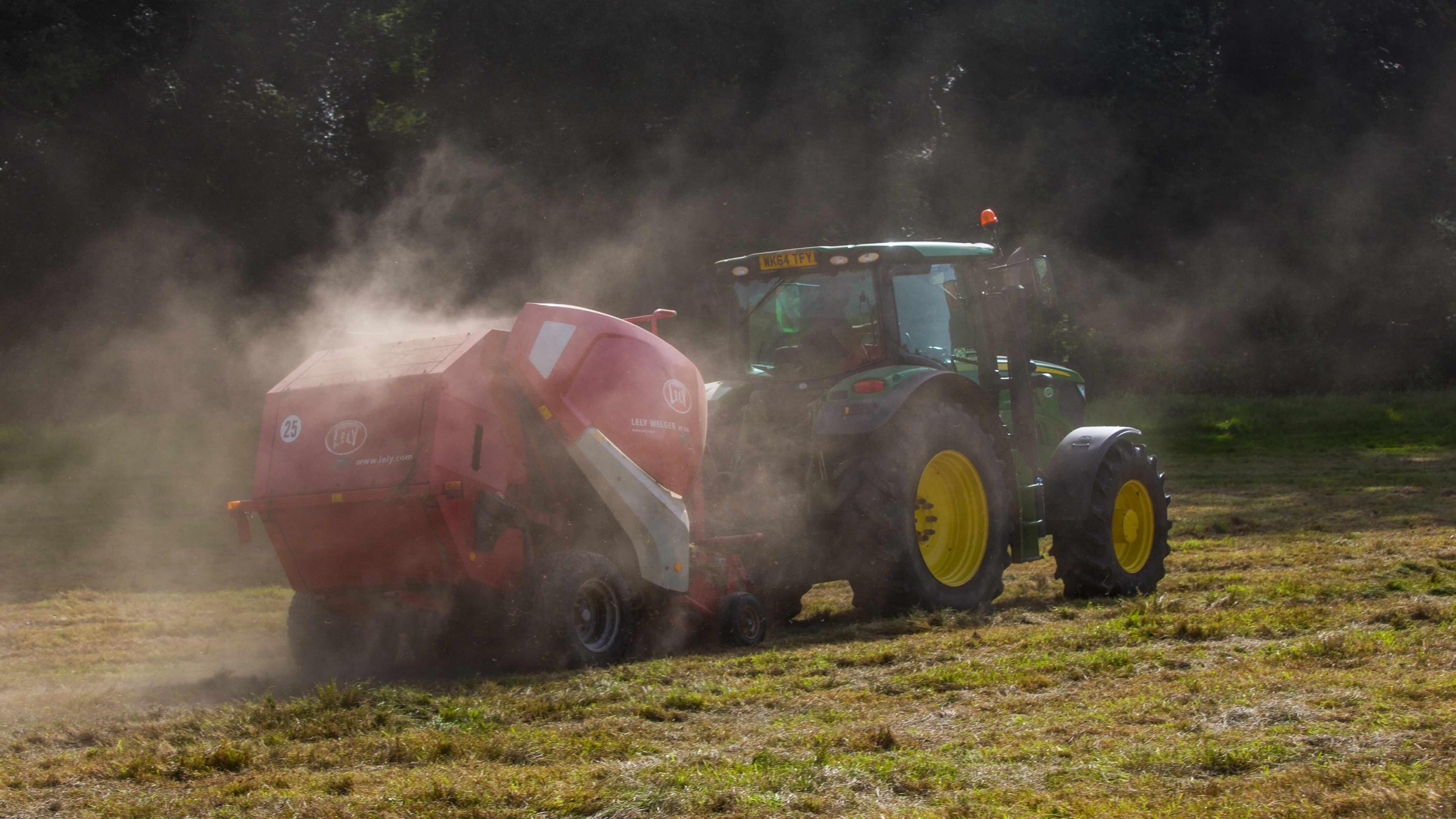 The image shows a green tractor with yellow wheels operating in a rural field, pulling a red baler—a machine used to collect and compress cut grass or hay into bales. Dust surrounds the machinery. The backdrop features trees and lush greenery.