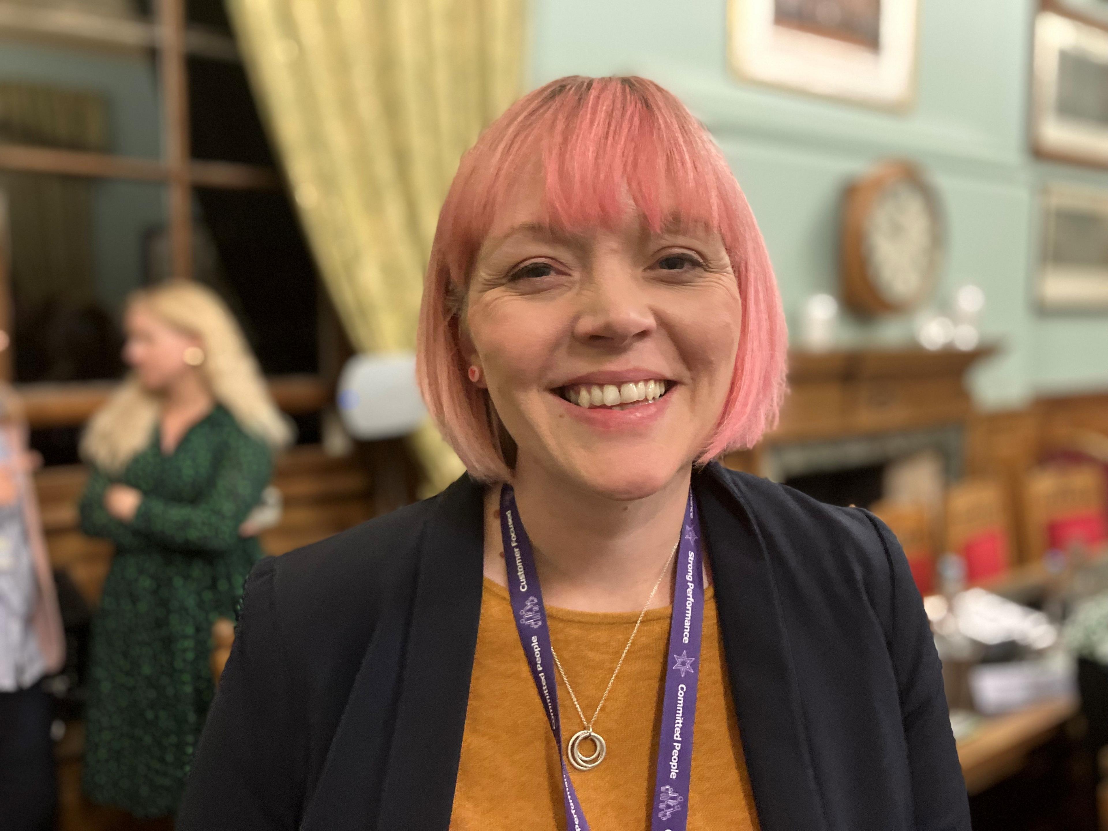 Katie Collier smiling at the camera in the council chamber. She has pink hair, a smart jacket and a purple lanyard.