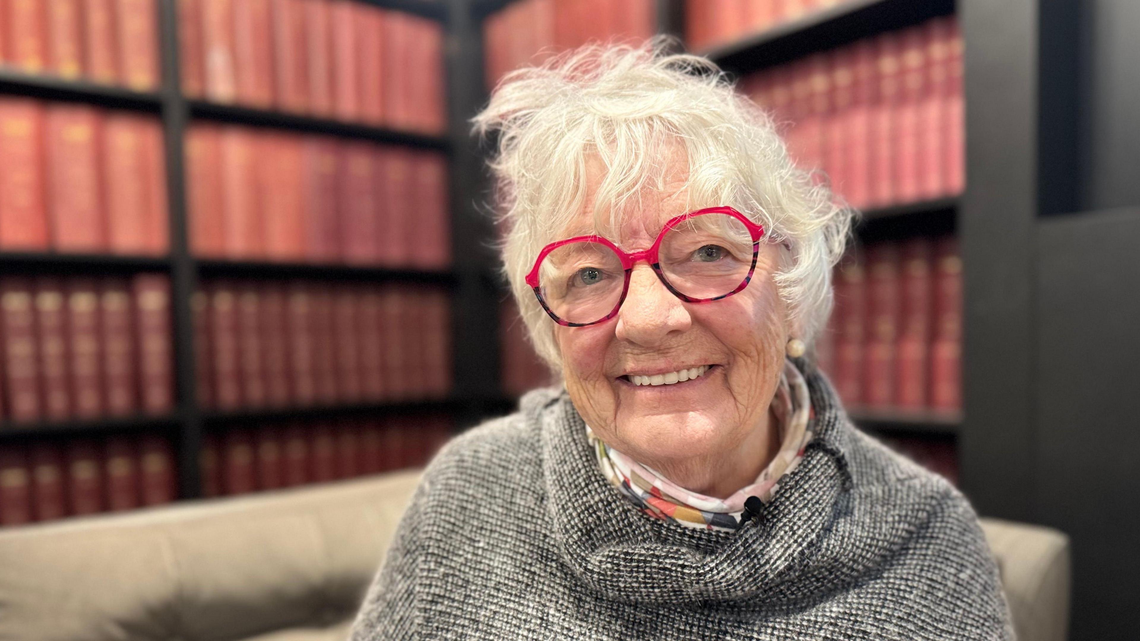 Yn y llun yma mae Viv yn gwenu. Mae ganddi siwmper llwyd ymlaen, gyda sgarff lliwgar. Mae ganddi wallt gwyn a sbectol coch.